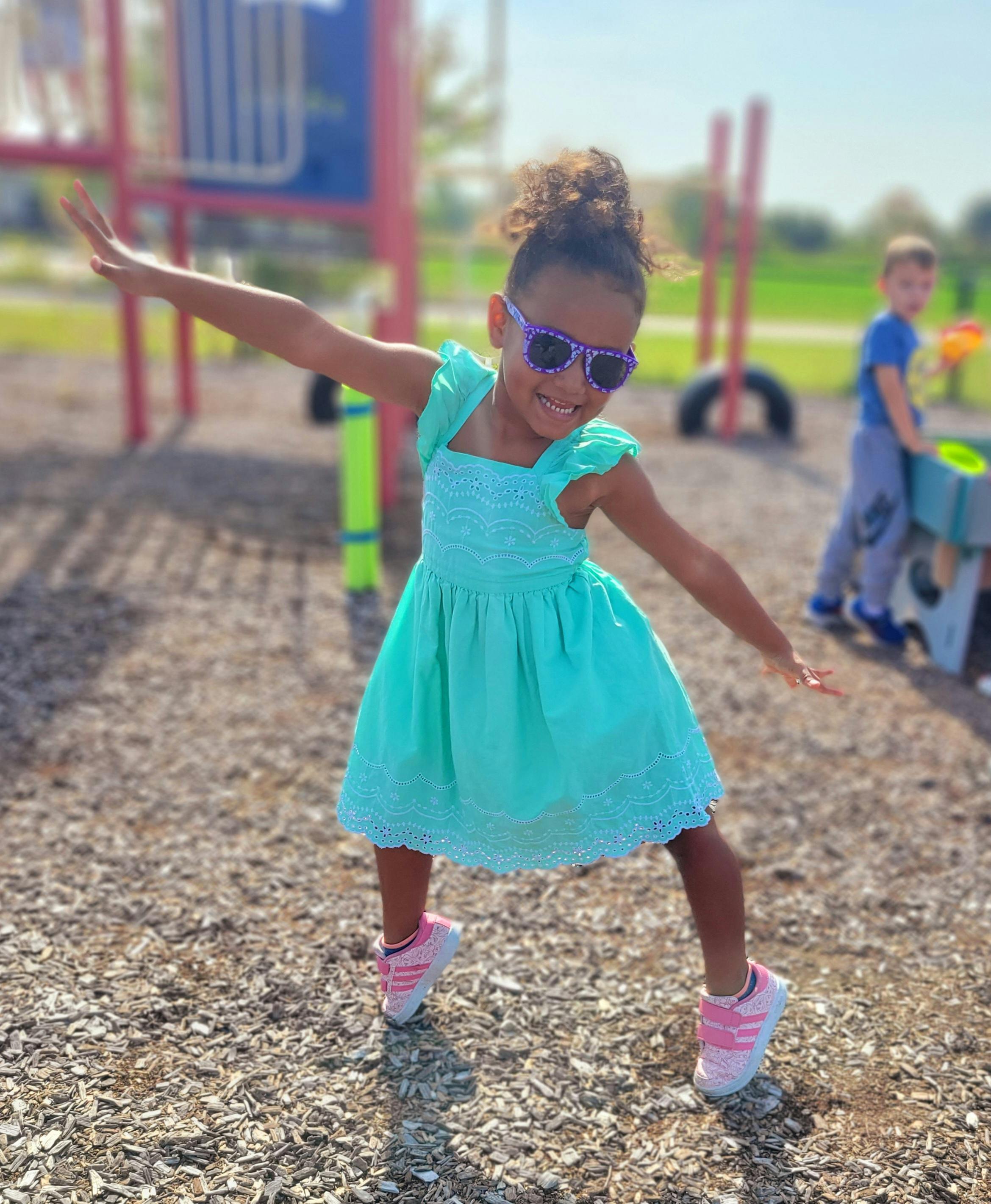 Child in sunglasses and dress with arms outstretched on playground