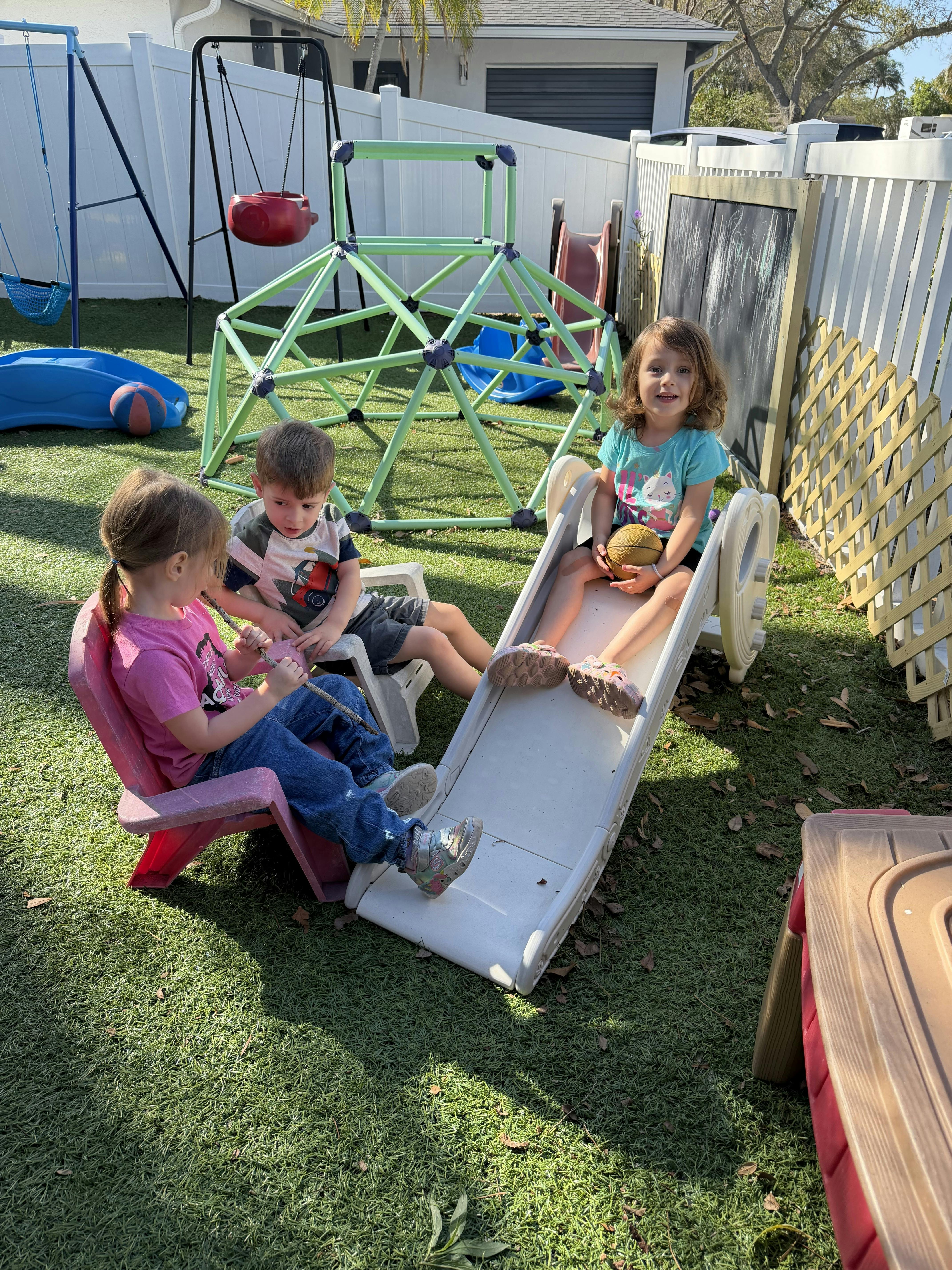 Three children playing outdoors, two on chairs and one on a slide holding a basketball