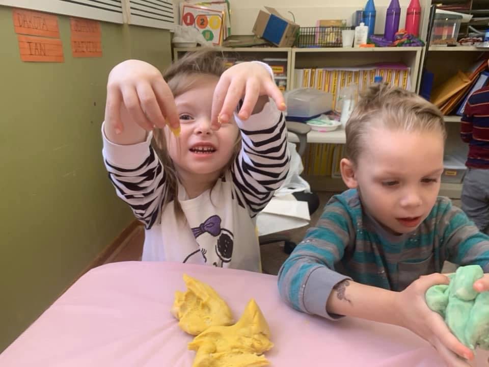 Child holding up small object, another child playing with modeling clay
