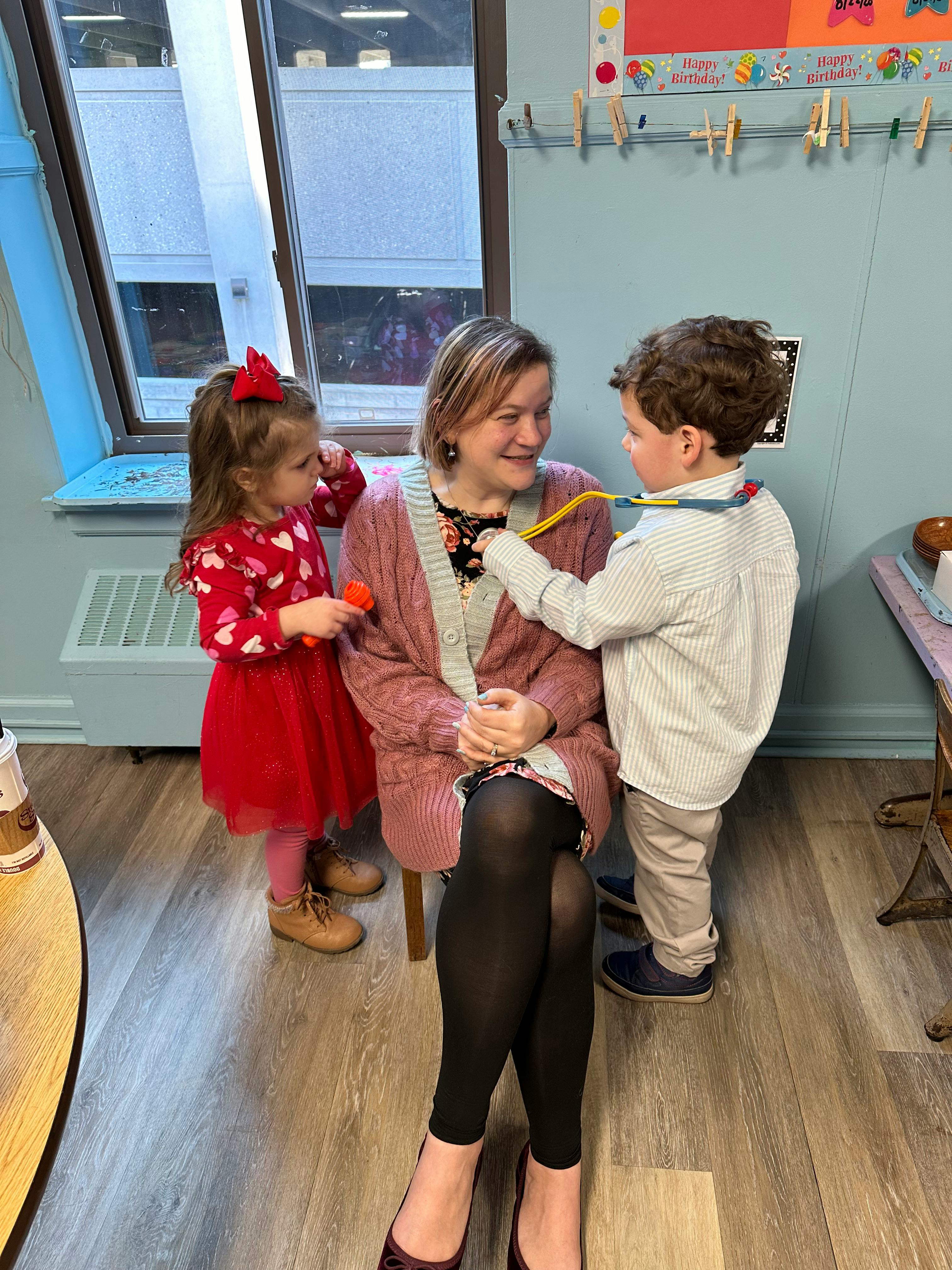 Person sitting with two children, one holding a toy and one playing with a toy stethoscope
