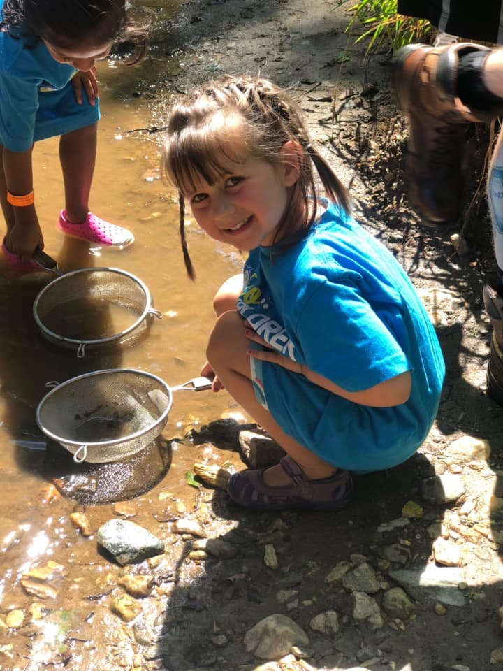 Child crouching in shallow water holding a strainer, another child nearby