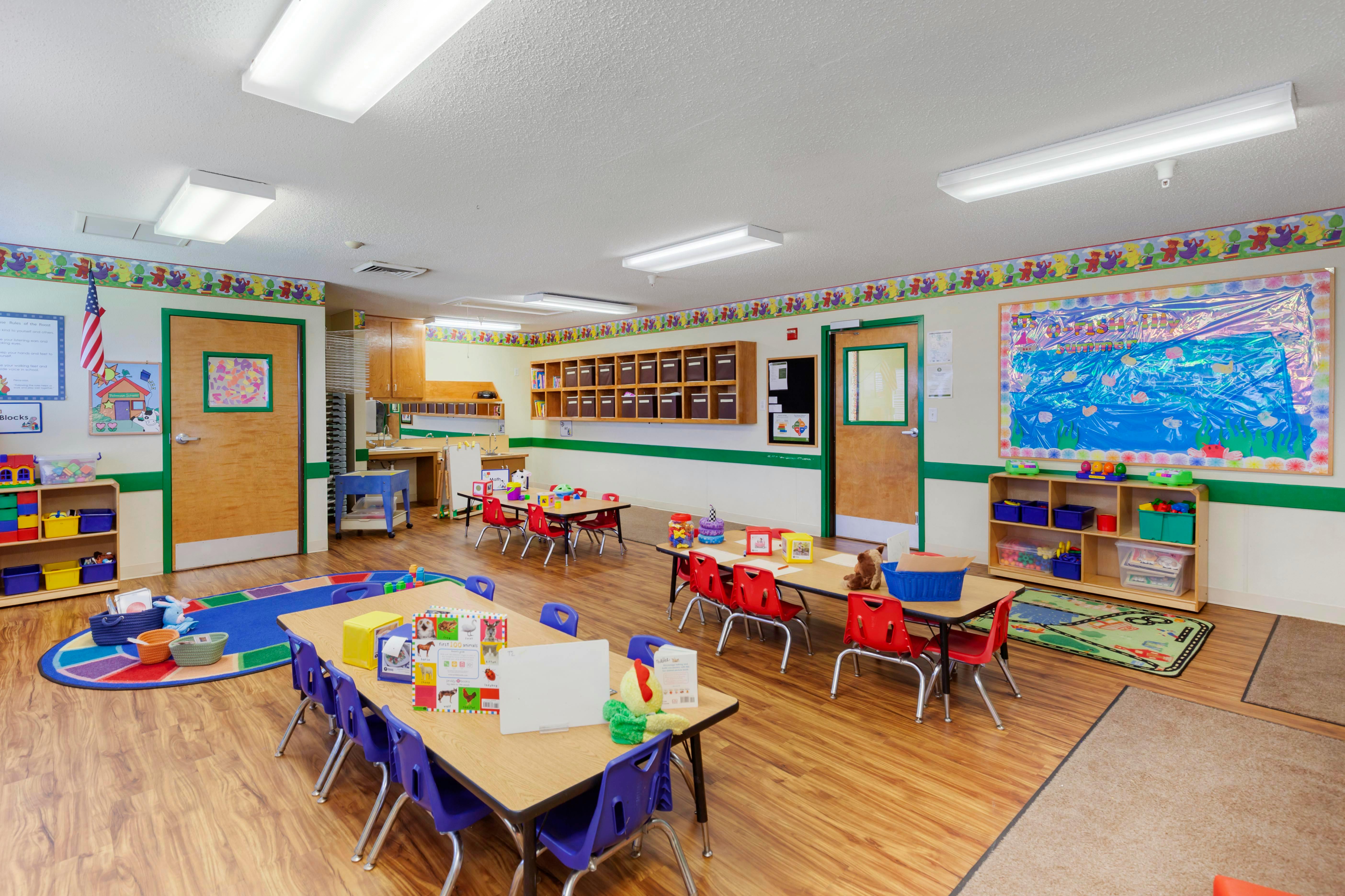 Classroom with small tables and chairs, shelves with toys, and bulletin boards