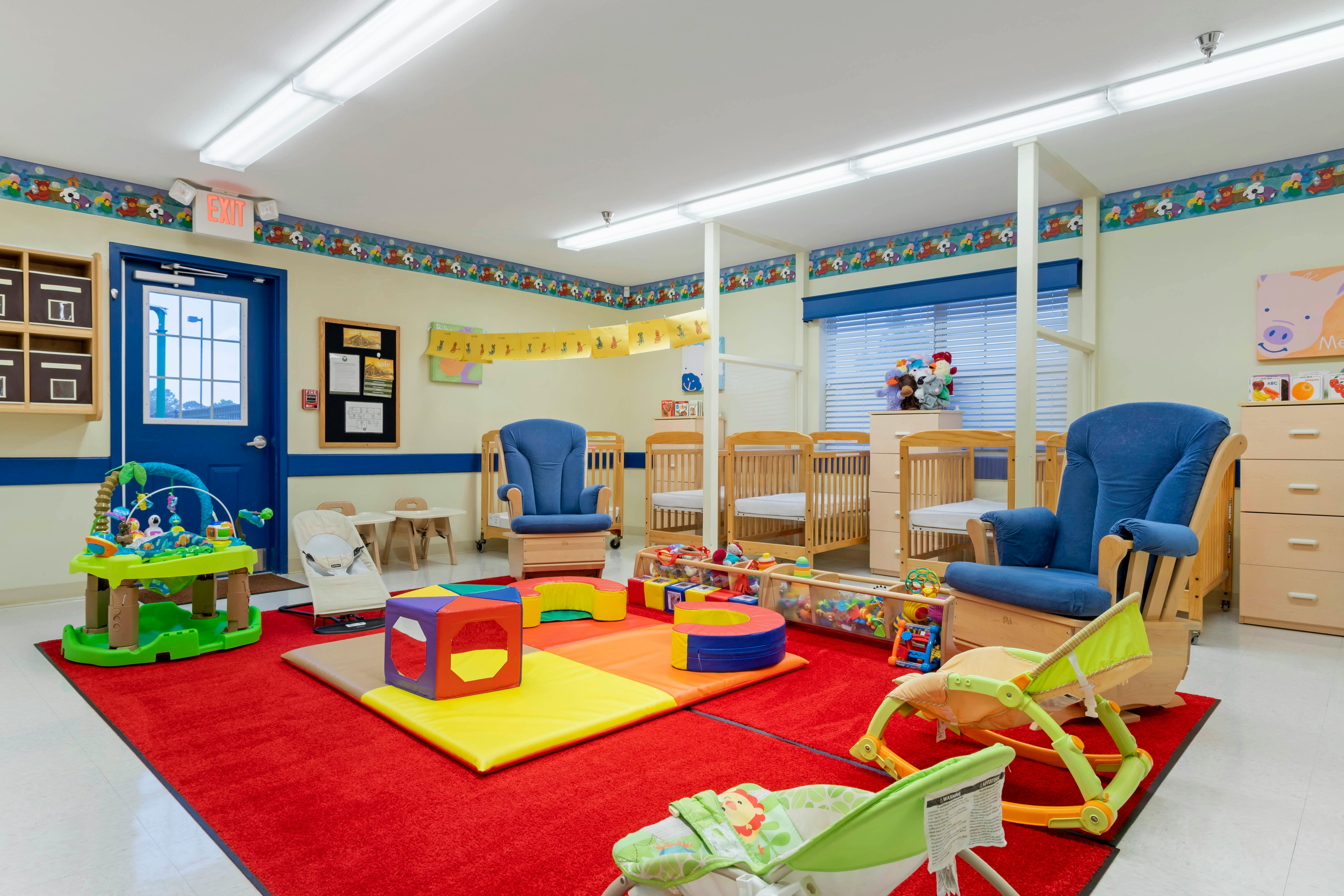 A colorful childcare room with cribs, play mats, toys, and blue rocking chairs