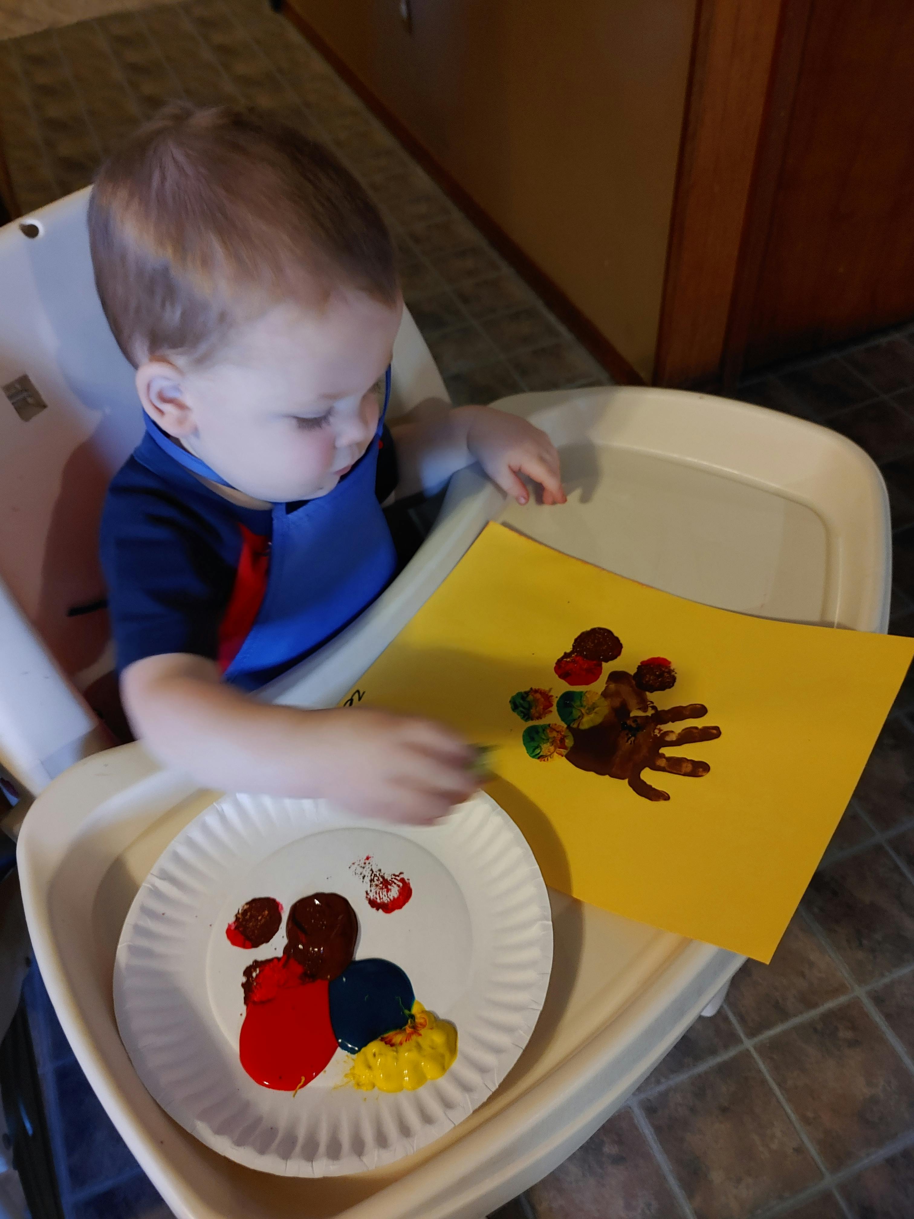 Child in a high chair painting a handprint turkey on yellow paper