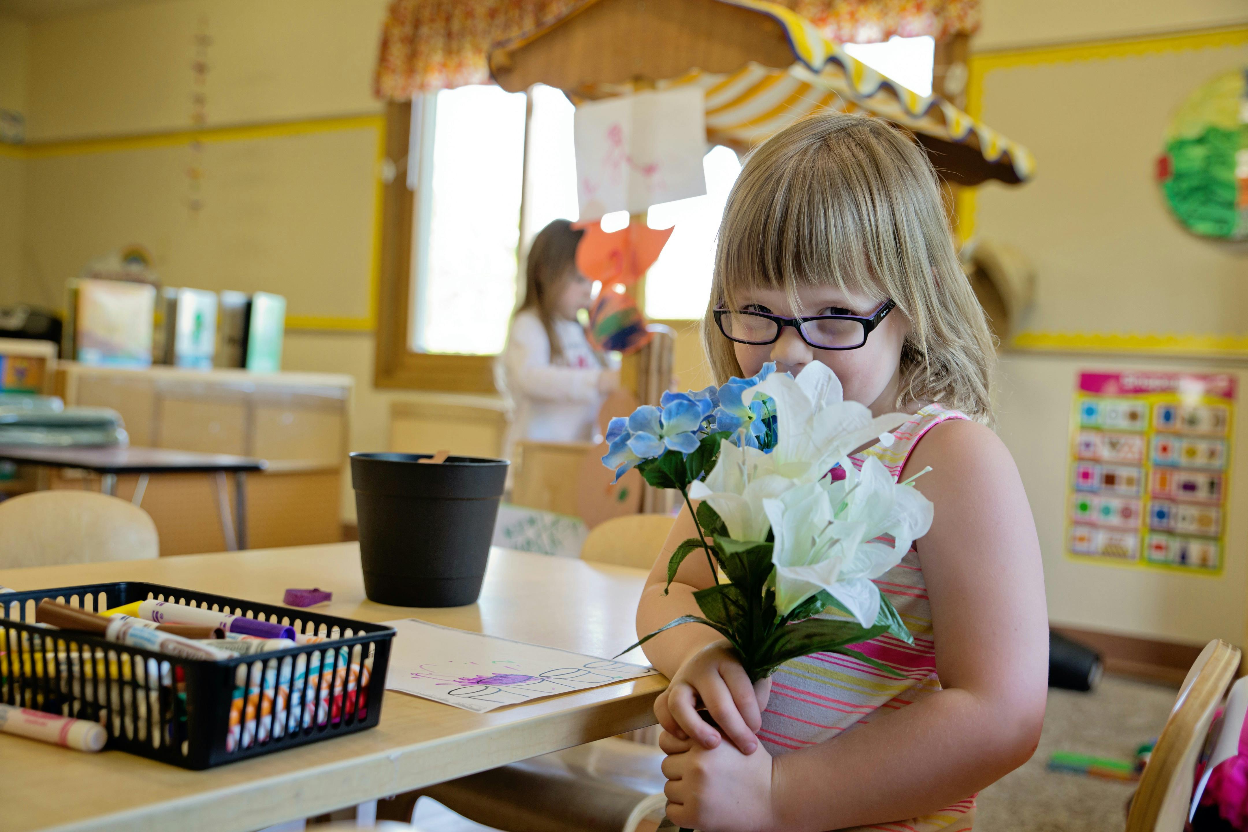 Child in glasses holding flowers in a classroom