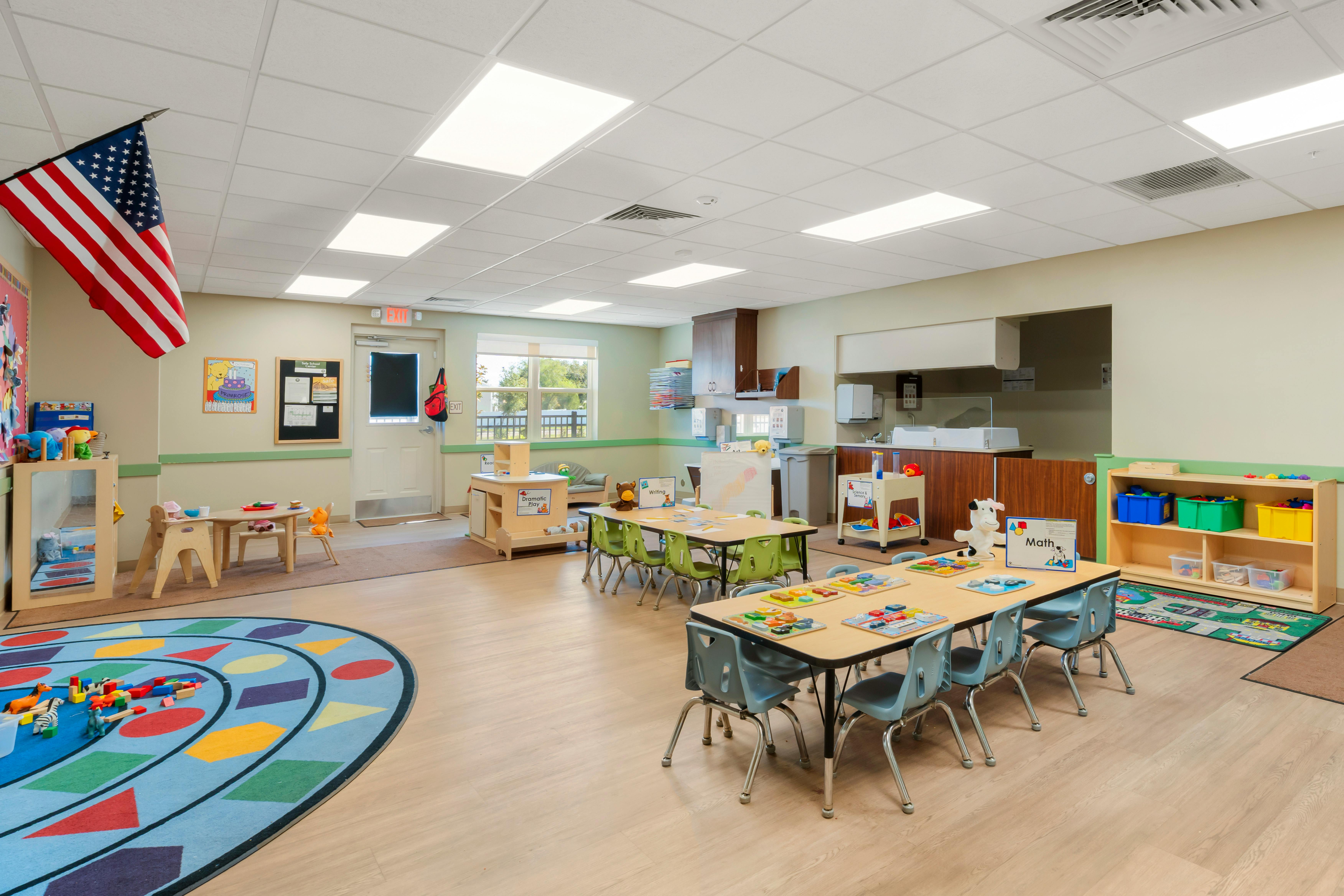 Classroom with multiple activity tables, a play rug, and an American flag