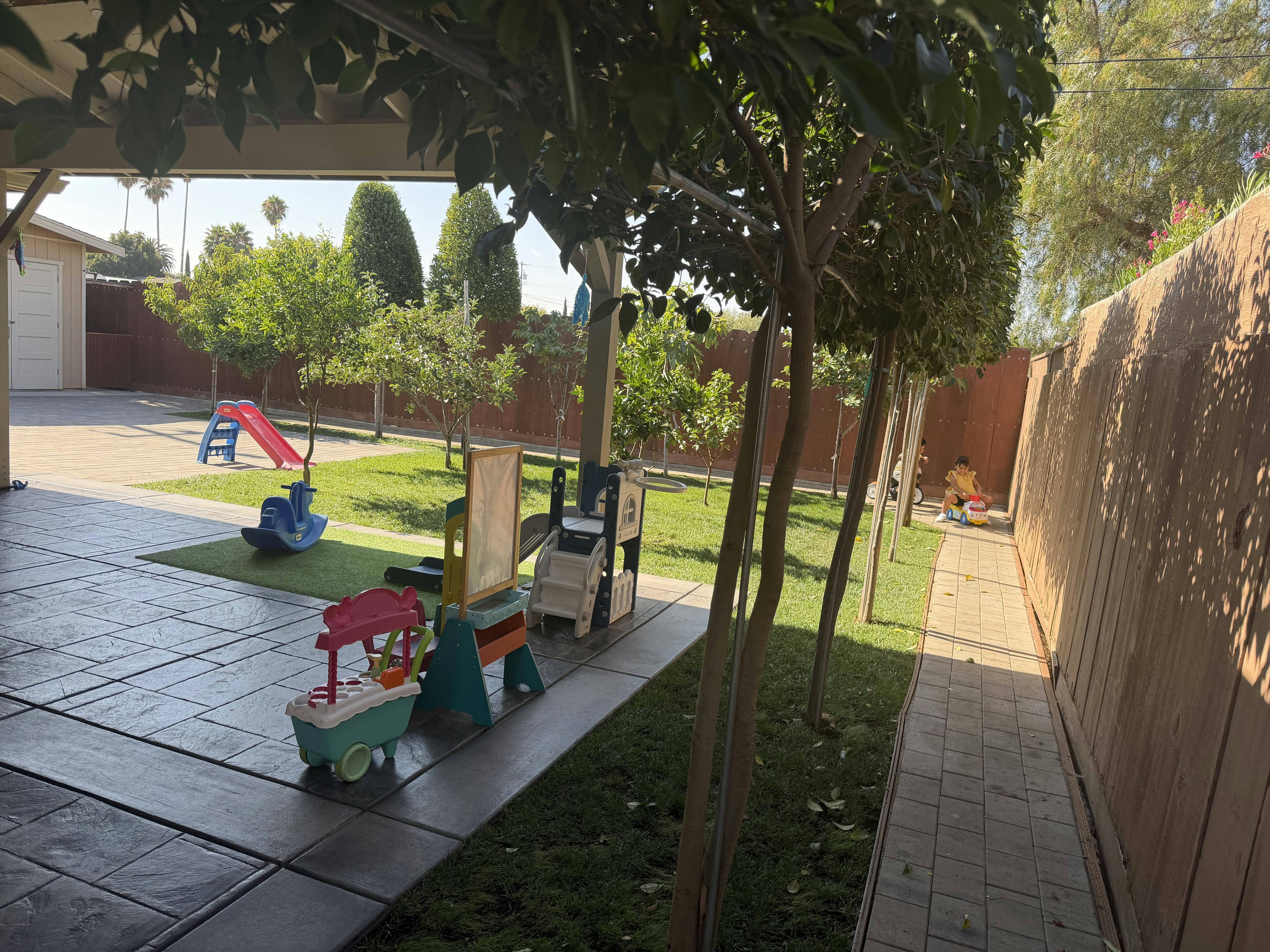 Child riding a toy car on a backyard patio with various play equipment