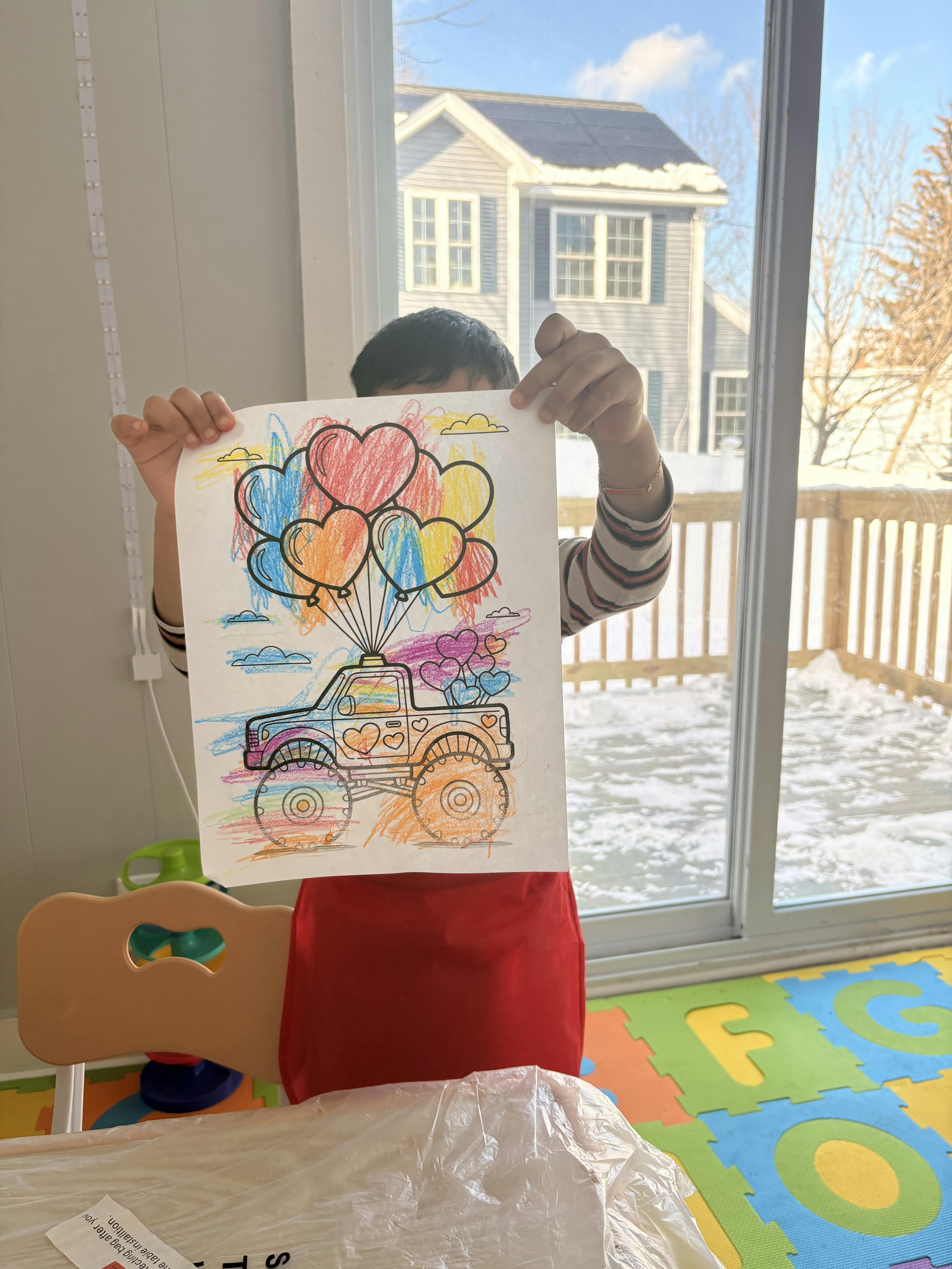 Child holding up a coloring page of a monster truck with heart-shaped balloons