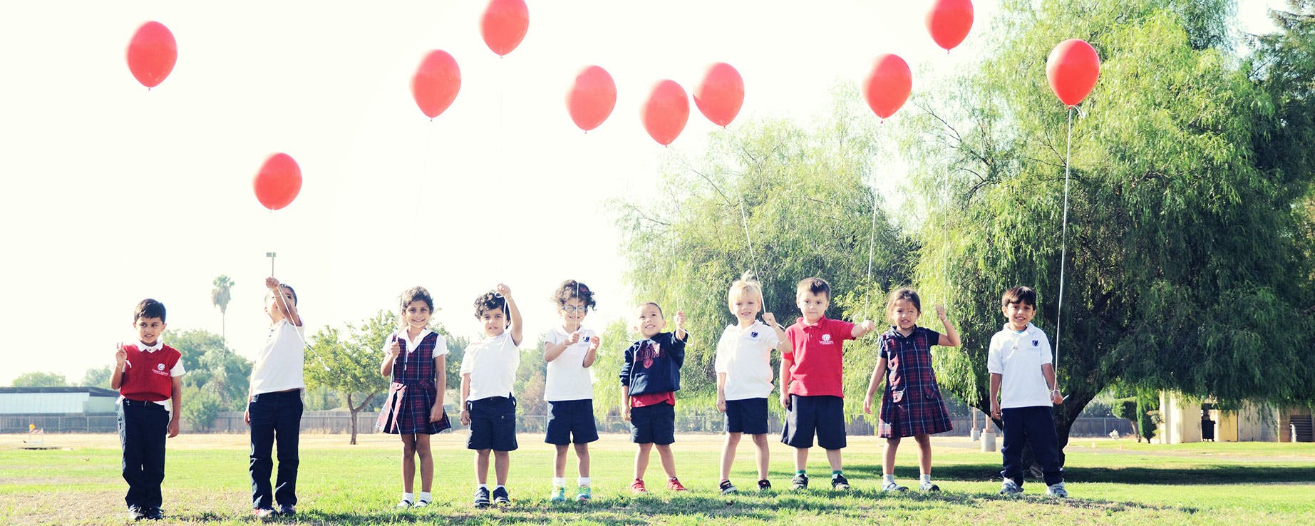 Children standing in a line outdoors with red balloons floating above them