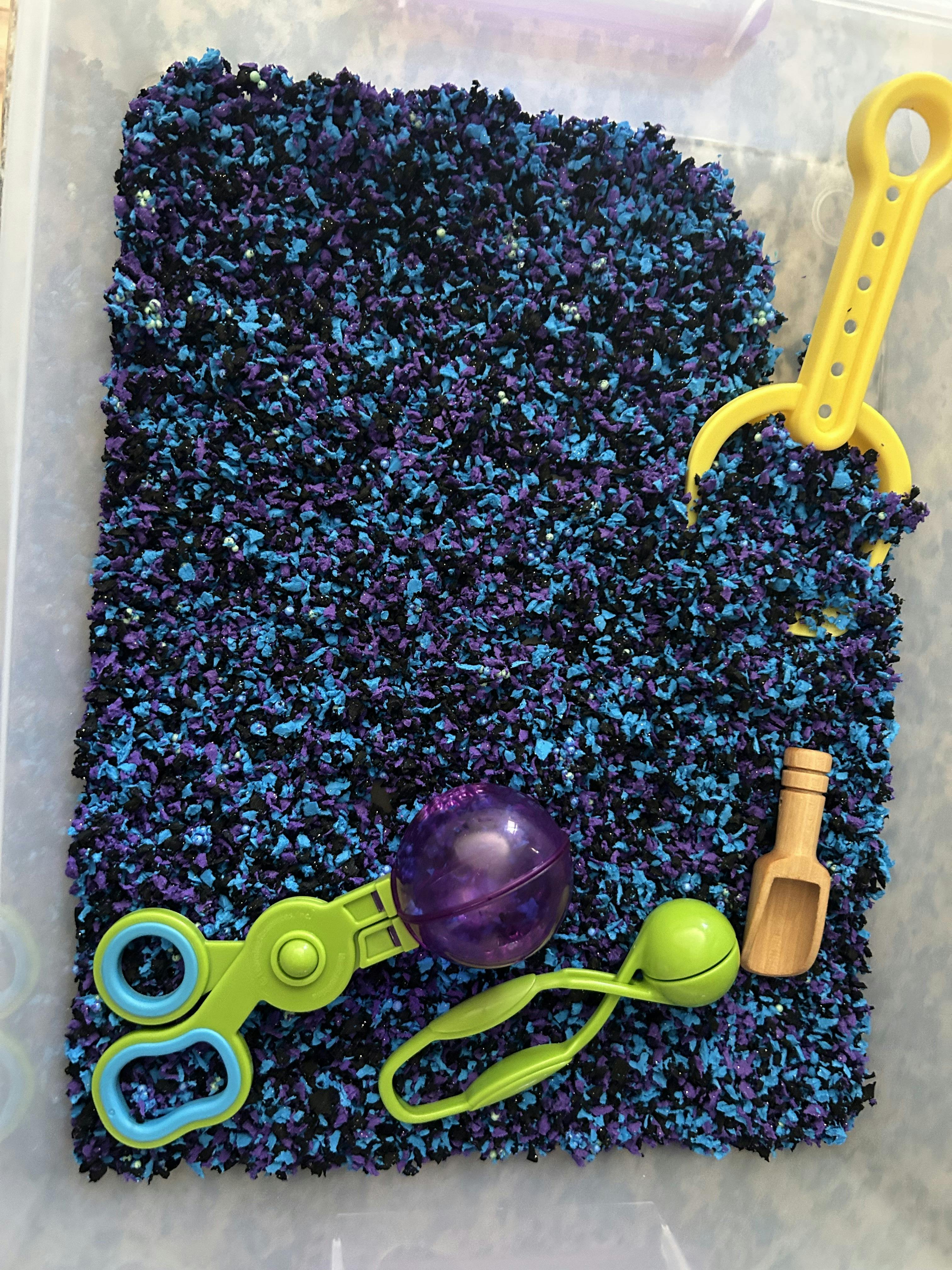 Sensory bin with blue and purple shredded paper, scoop, and plastic toys