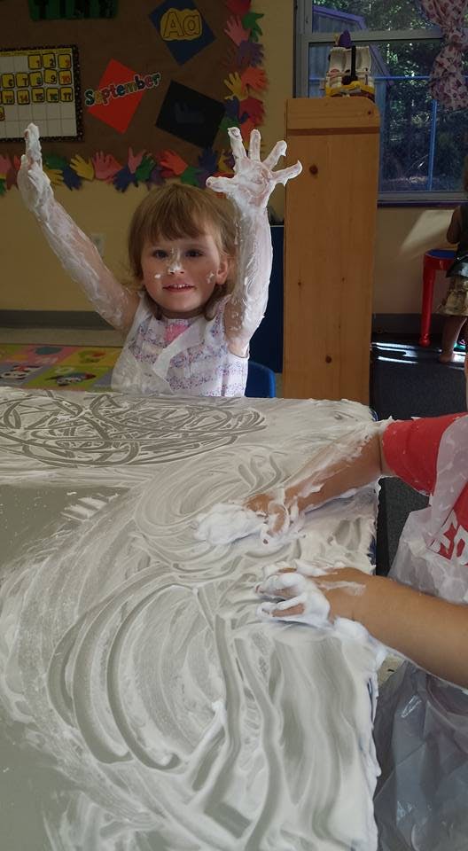 Child with arms raised, covered in white foam, with another child playing in a classroom
