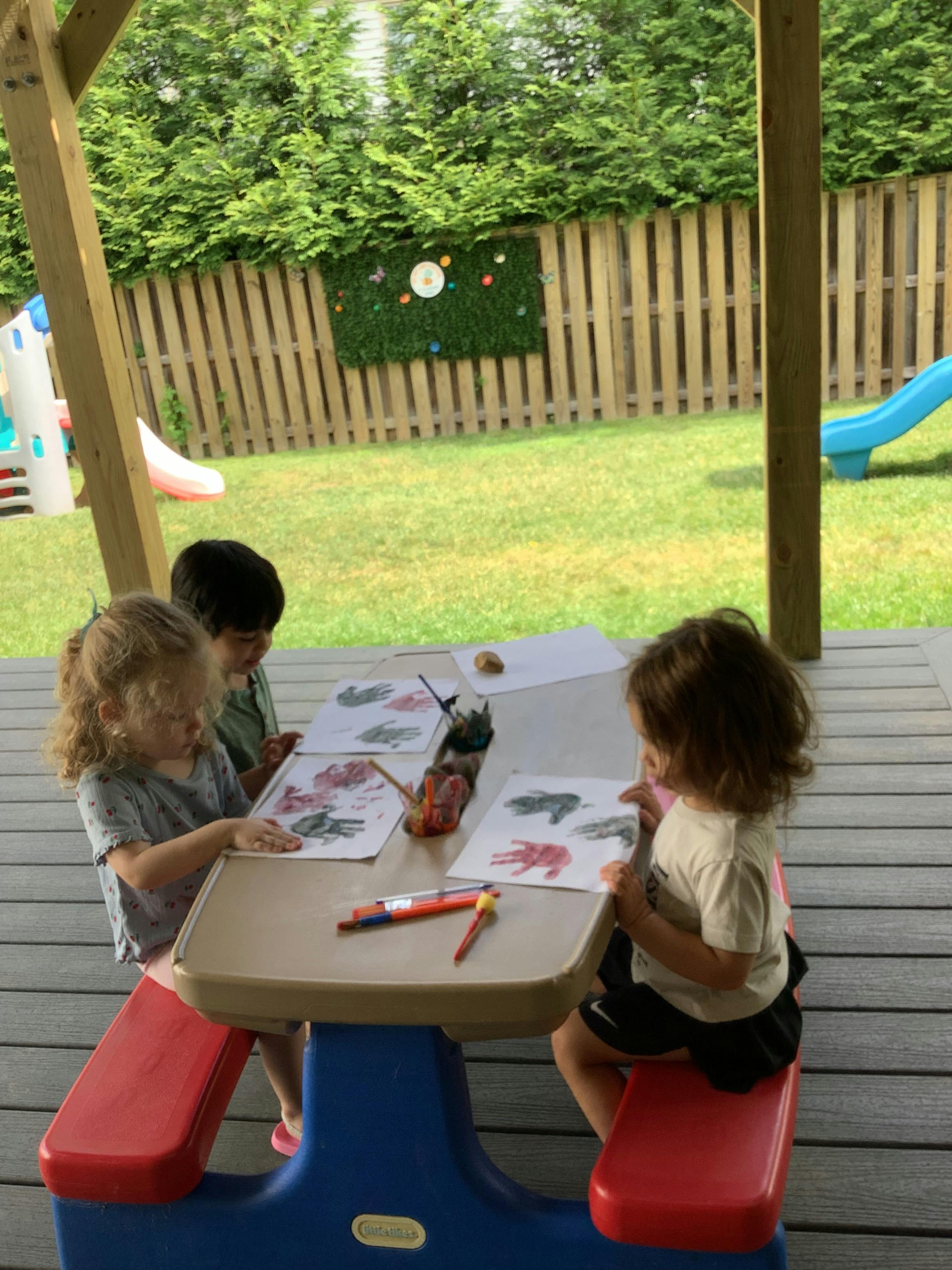 Children painting handprints on paper at a small table outdoors