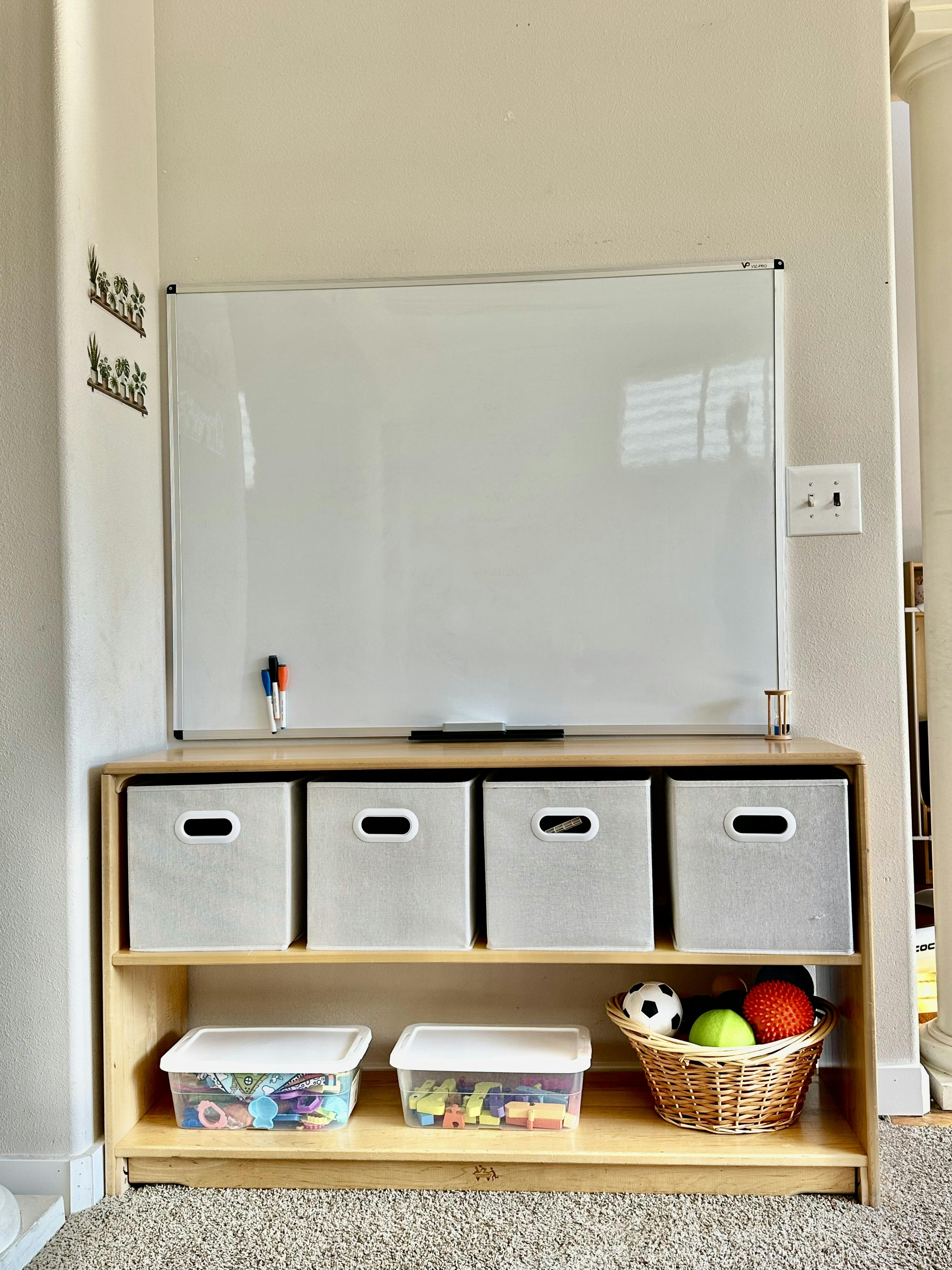 Whiteboard mounted above a wooden shelving unit with storage bins and toys