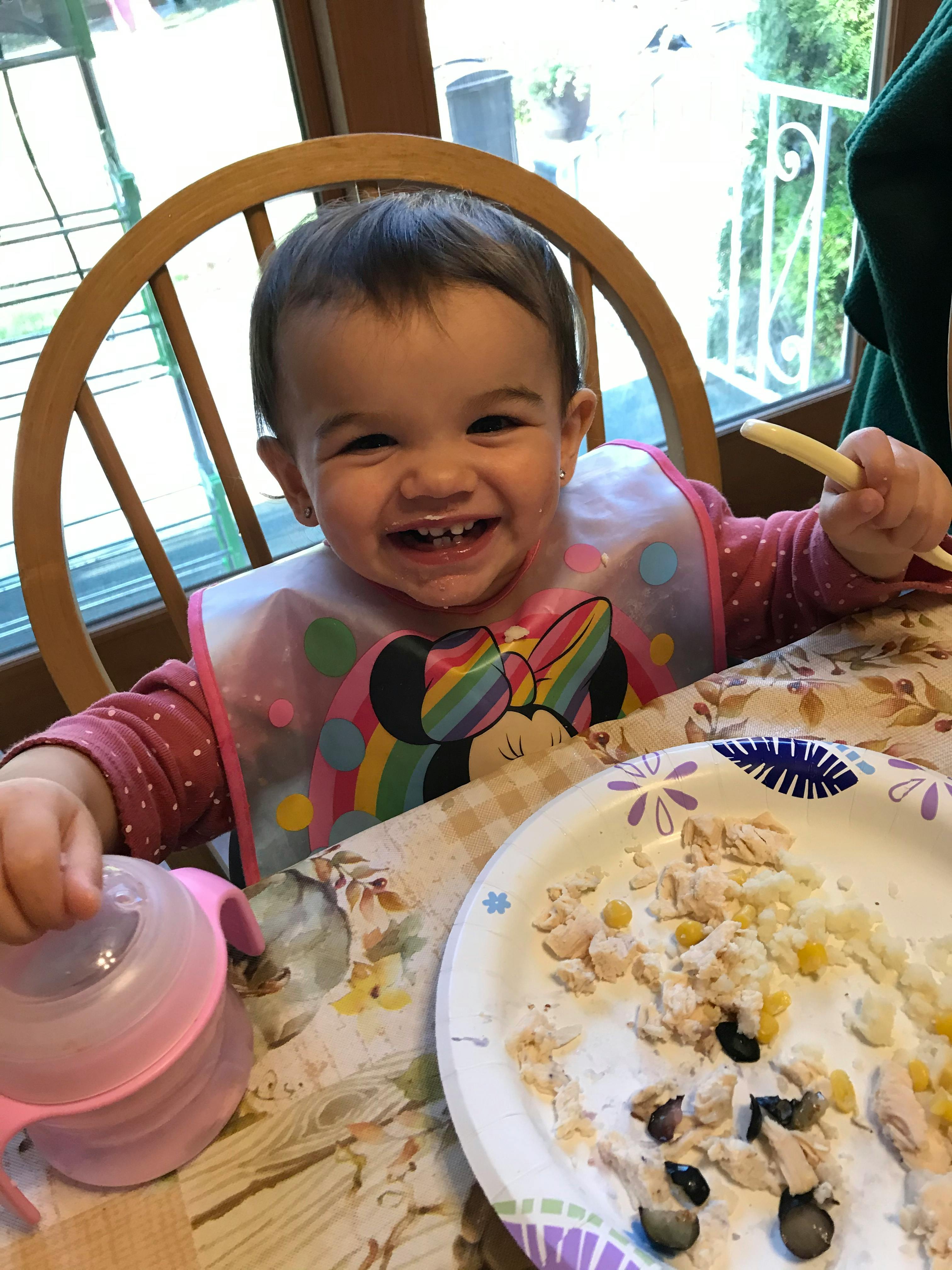 Child in a high chair with a bib, holding a spoon and a sippy cup