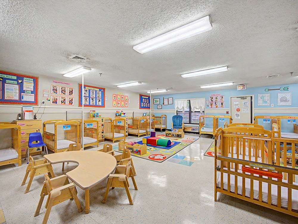 daycare classroom with multiple cribs, a U-shaped table with chairs, and a playmat