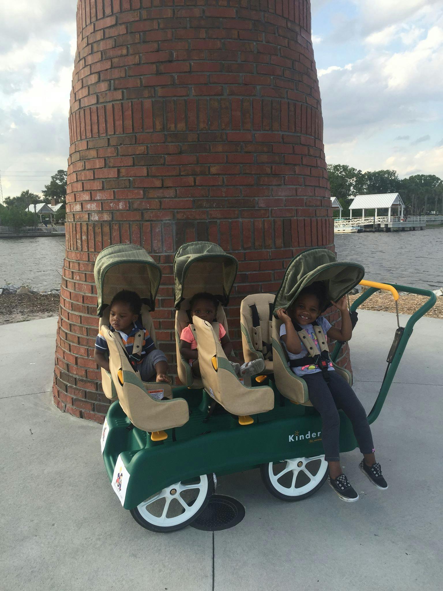 Three children in a multi-seat stroller next to a brick column