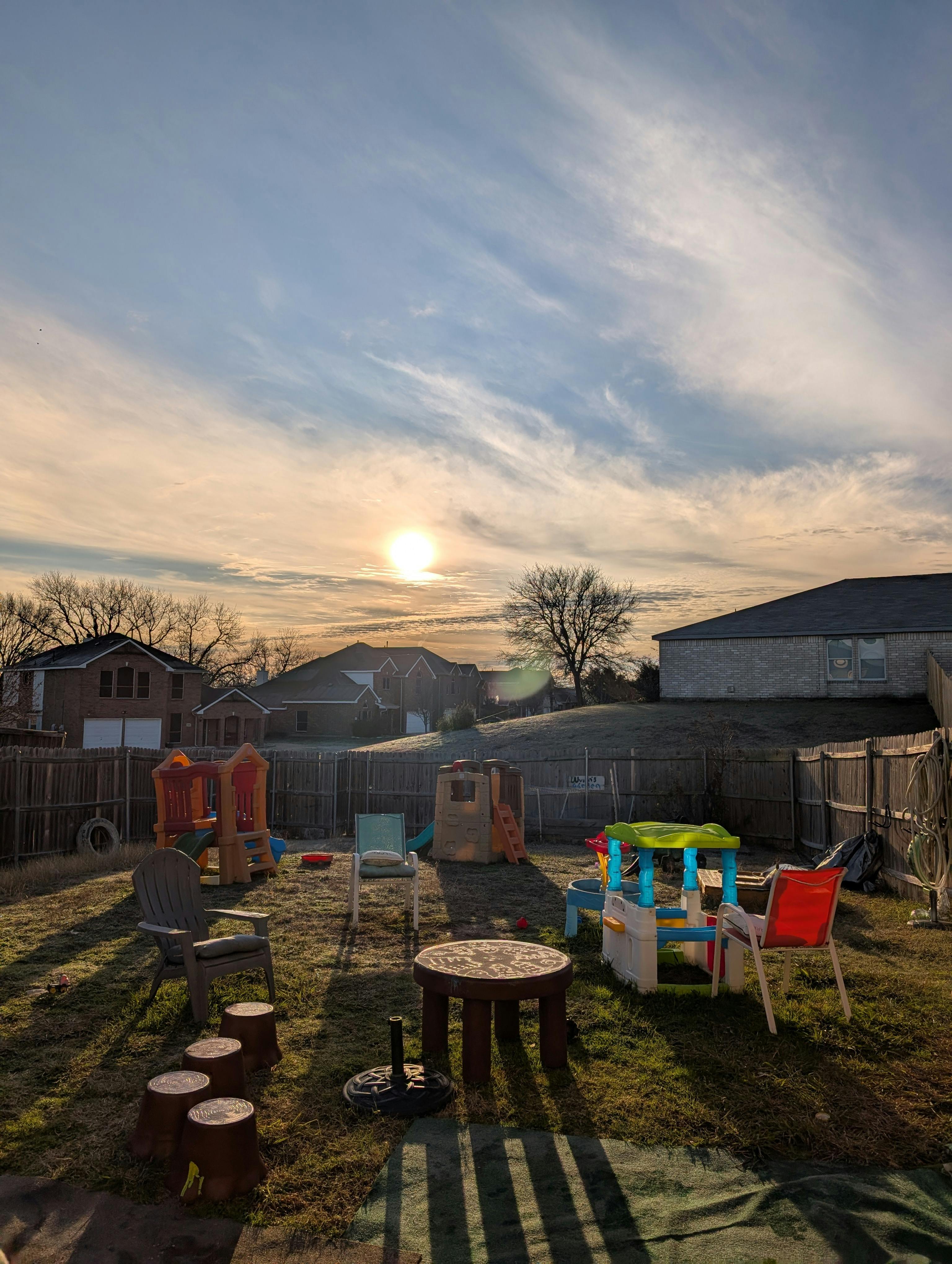 Backyard with play structures, chairs, and houses in the distance under a bright sky
