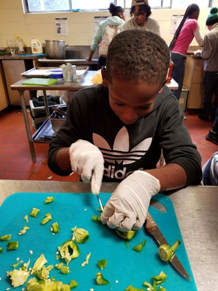 Child wearing gloves chopping green peppers on a blue cutting board in a kitchen