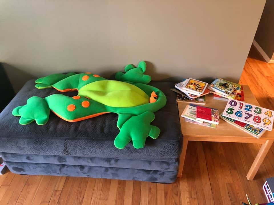 Green frog plush toy on a gray cushion next to a small table with books and a number puzzle
