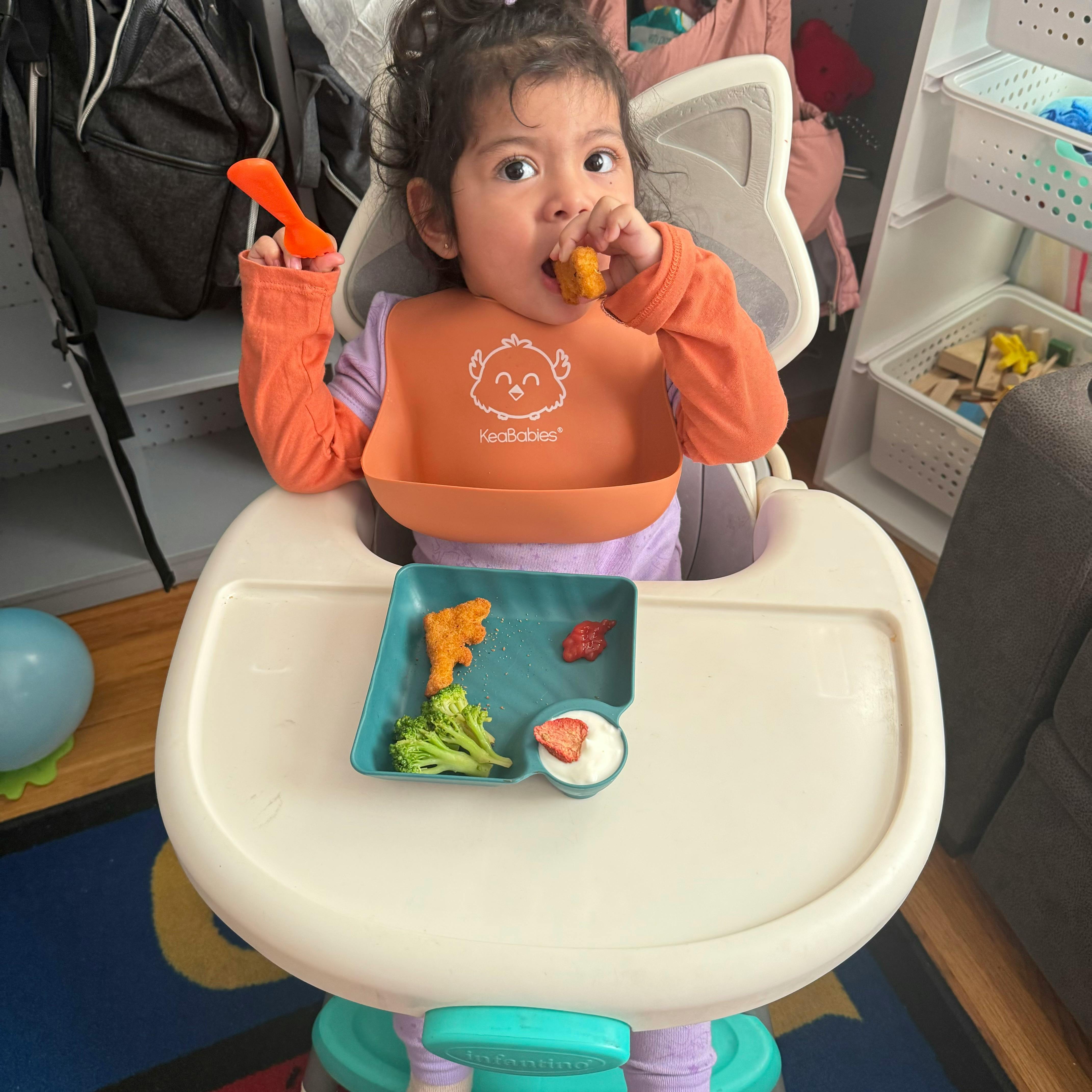 Child in a high chair eating food from a plate with broccoli and other food