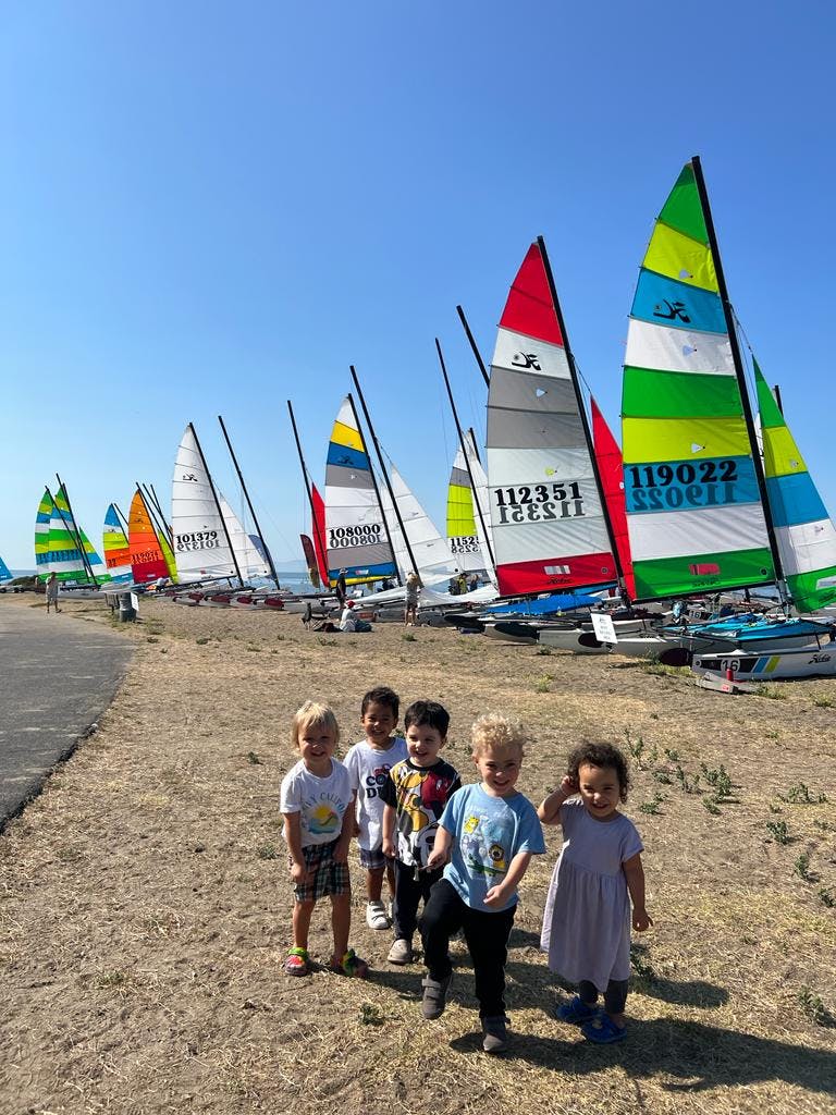 Five children standing on a beach with sailboats in the background
