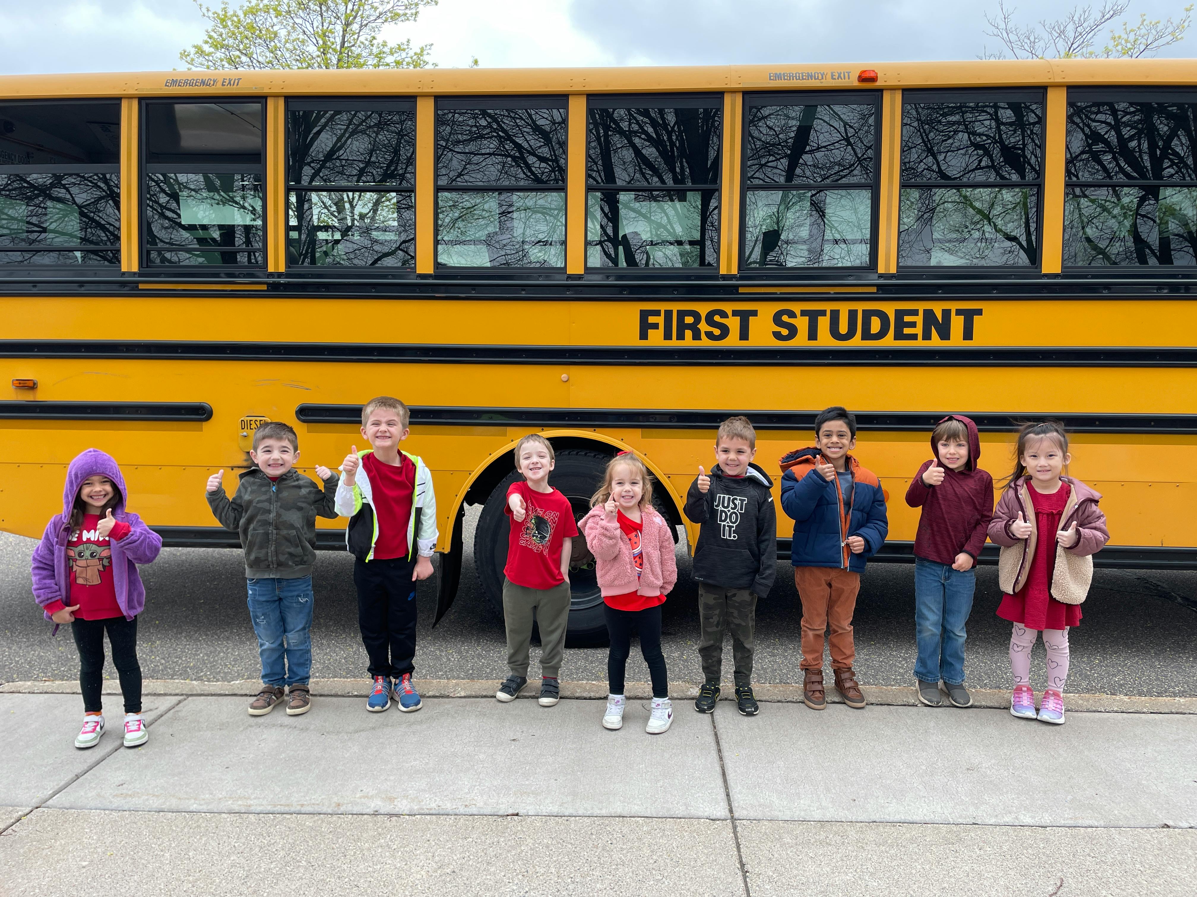 Group of children standing in front of a school bus with "FIRST STUDENT" on the side