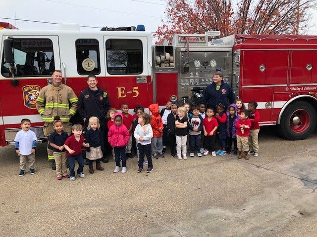Two people in firefighter uniforms with a group of children in front of a fire truck