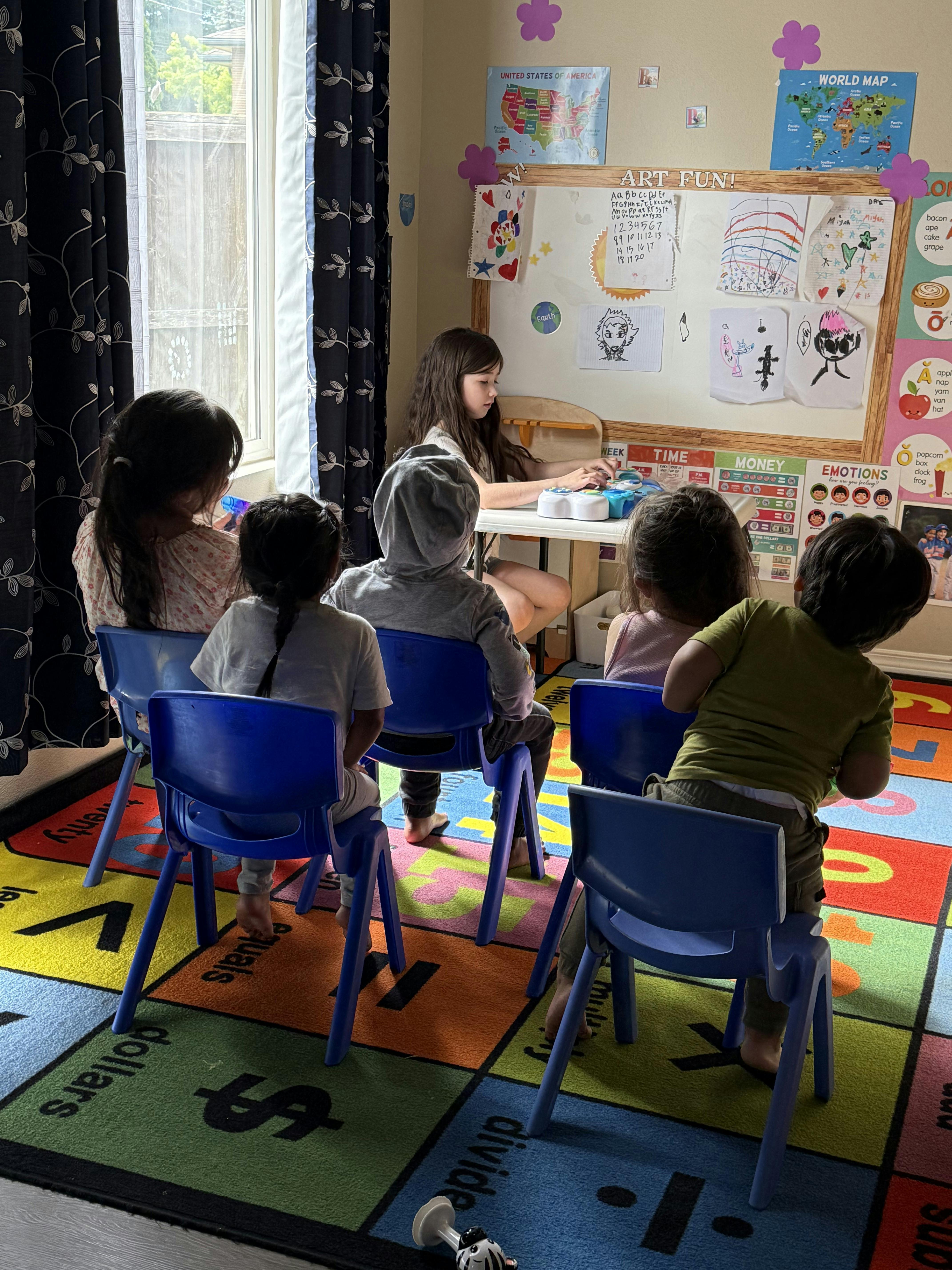 Children sitting on blue chairs in a classroom, facing a person at a table