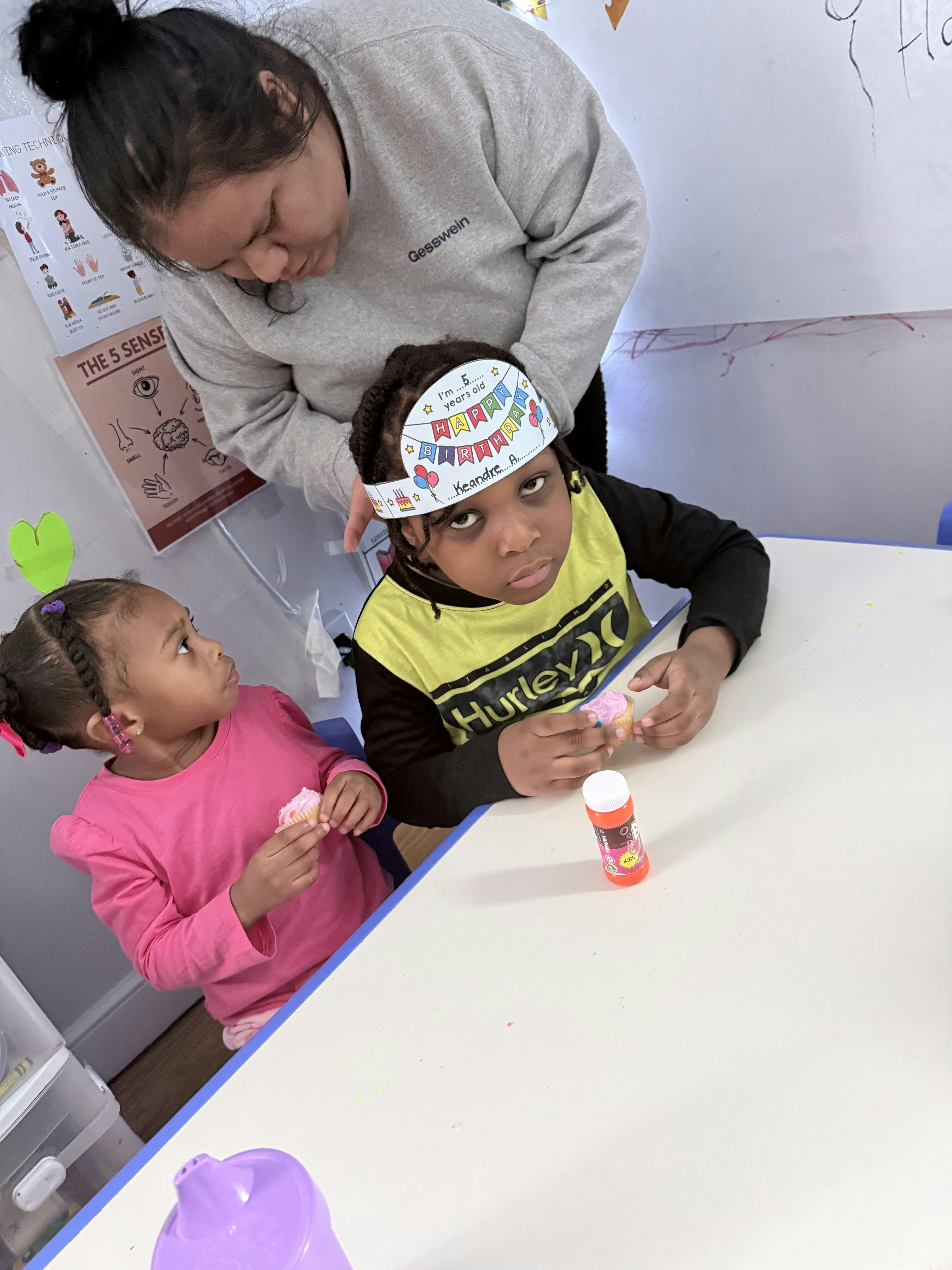 Adult helping a child wearing a birthday crown, another child holding a cupcake
