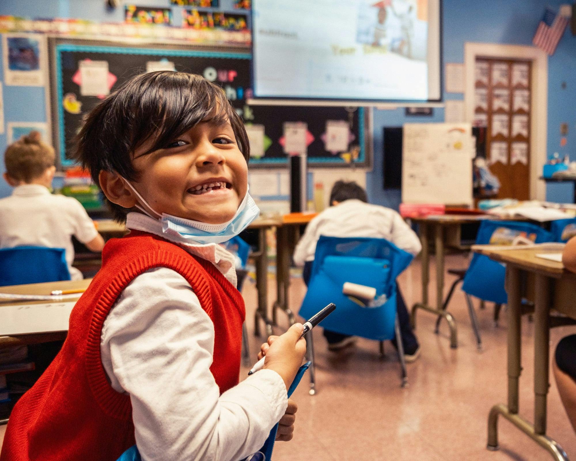Child in a red vest and white shirt smiling at the camera in a classroom