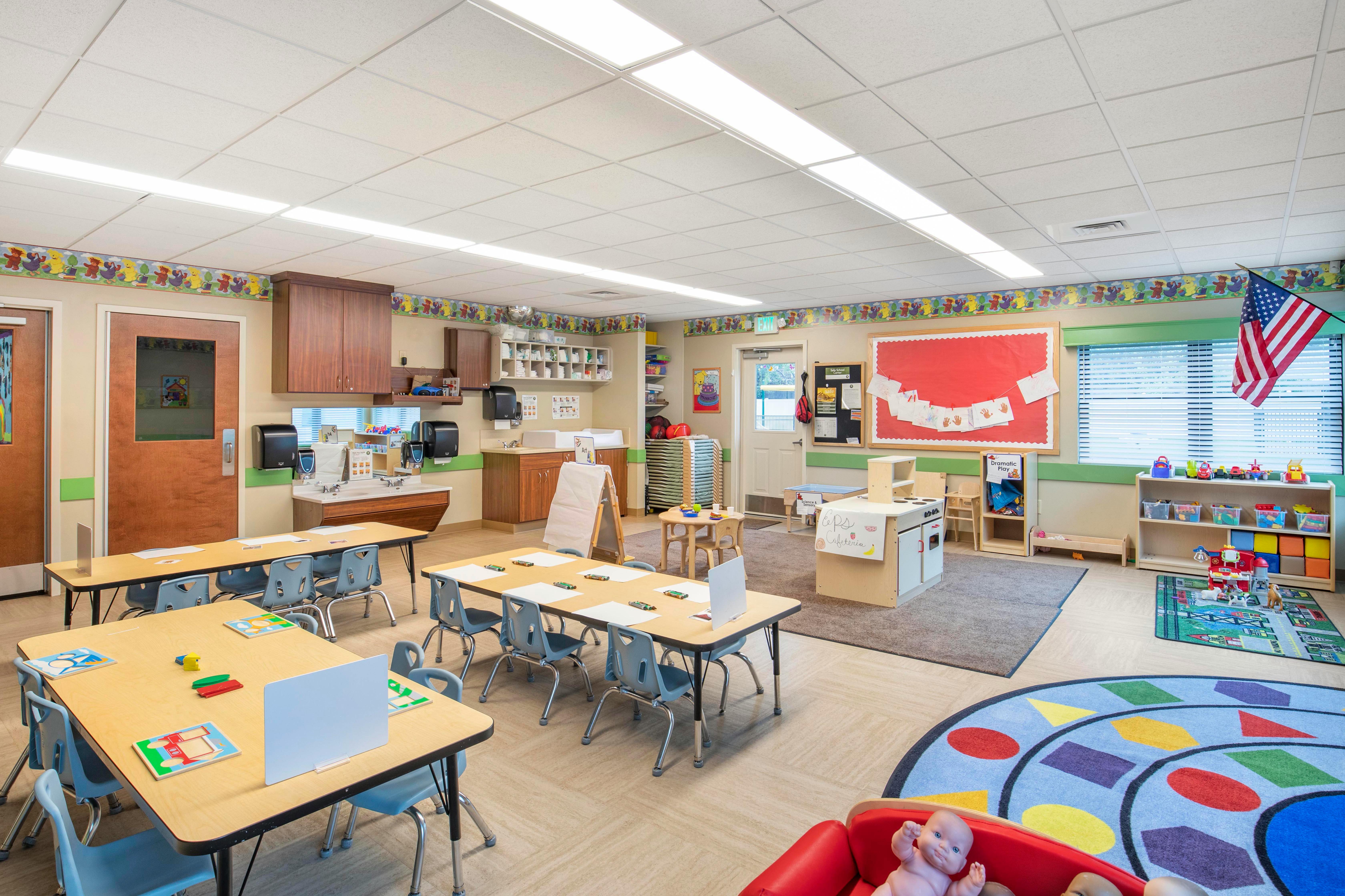 Classroom with tables and chairs, a play kitchen, and a circular rug with shapes