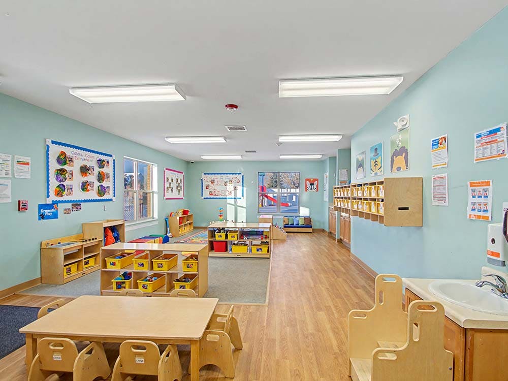 Classroom with small tables and chairs, shelves with yellow bins, and a sink