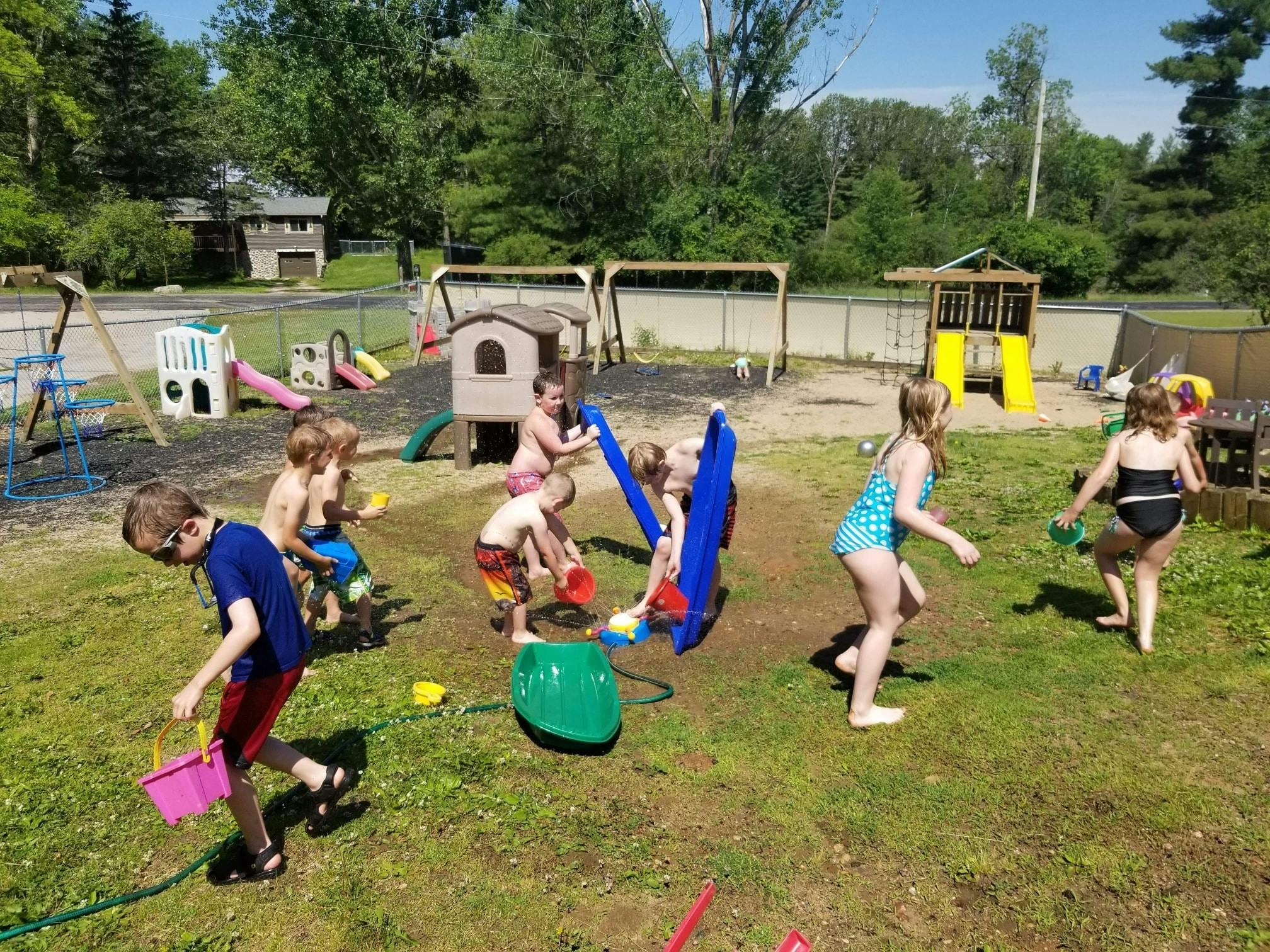 Children playing with water toys in a grassy area with a playground in the background