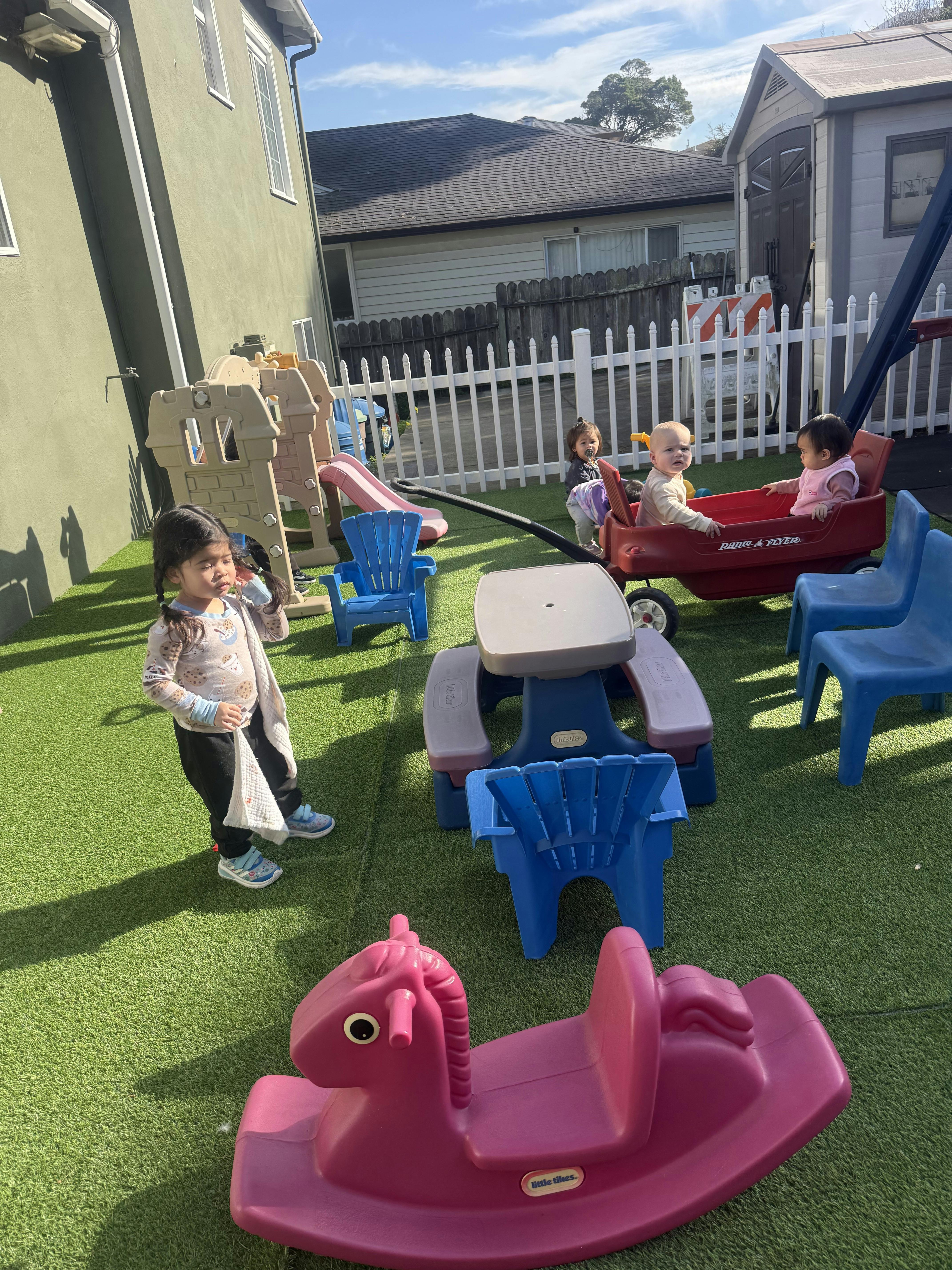 Child standing on artificial grass, with other children in a wagon and playground equipment