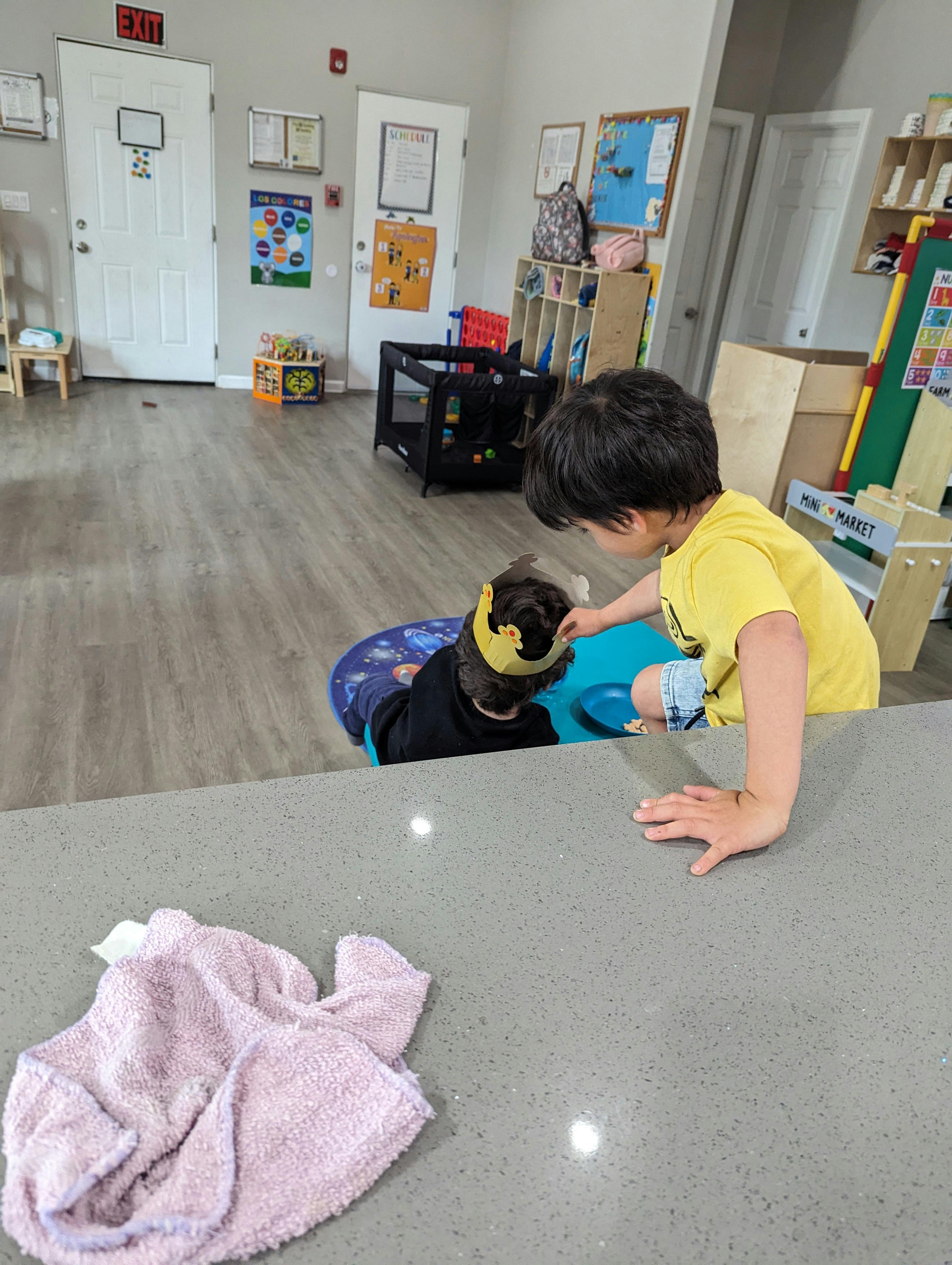 Child in yellow shirt reaching toward another child in a high chair