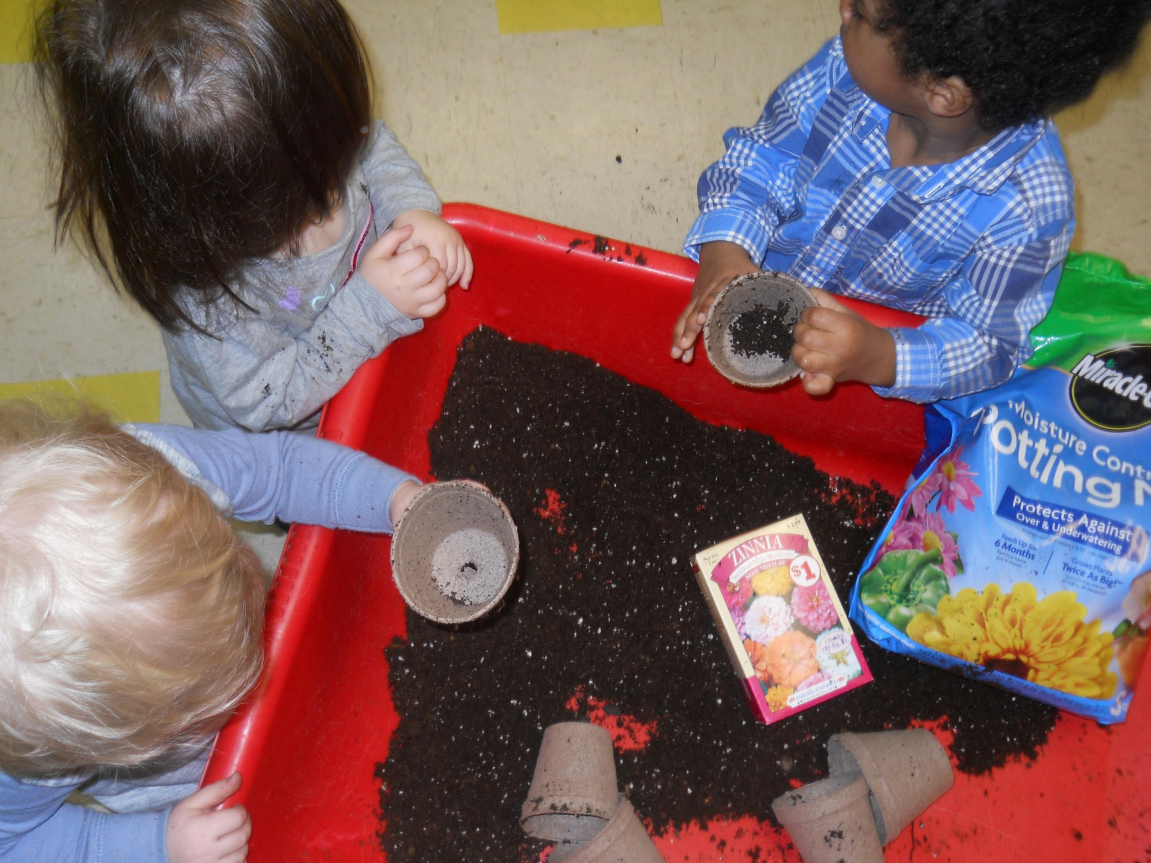 Three children playing with soil and small pots in a red bin