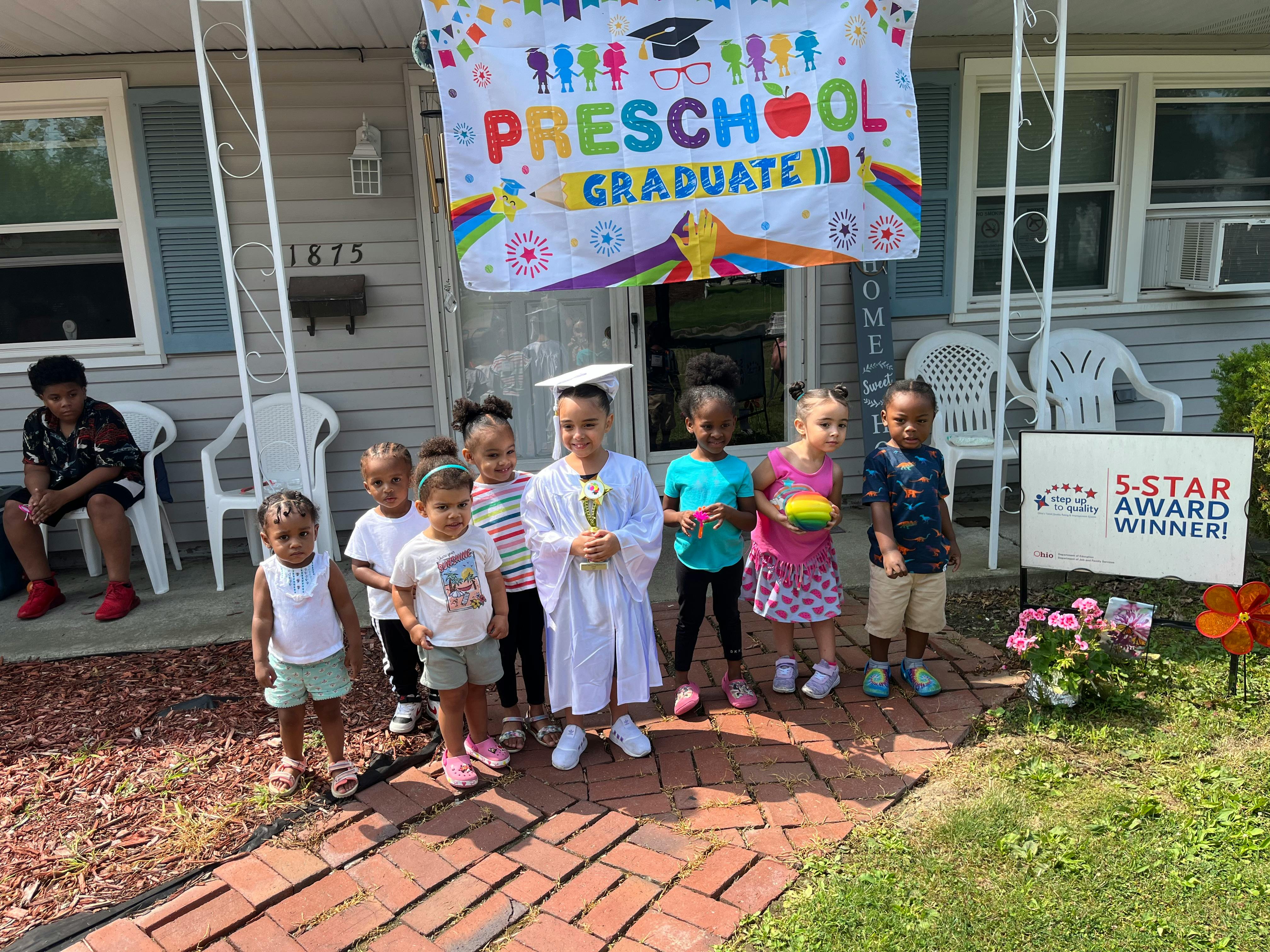 Children standing in front of a house with a "Preschool Graduate" banner
