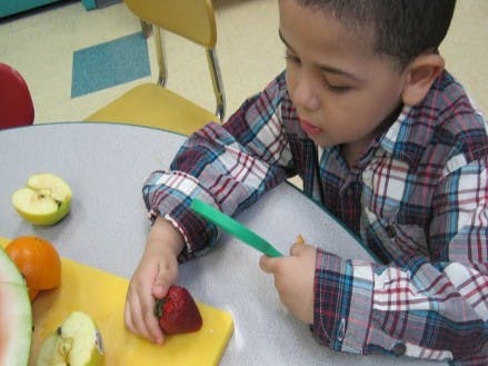 Child cutting a strawberry on a yellow cutting board with a green knife