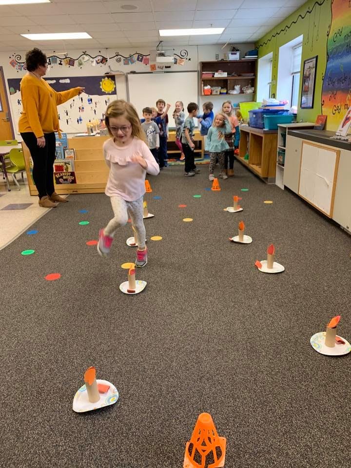 Child running through an obstacle course in a classroom with an adult and other children