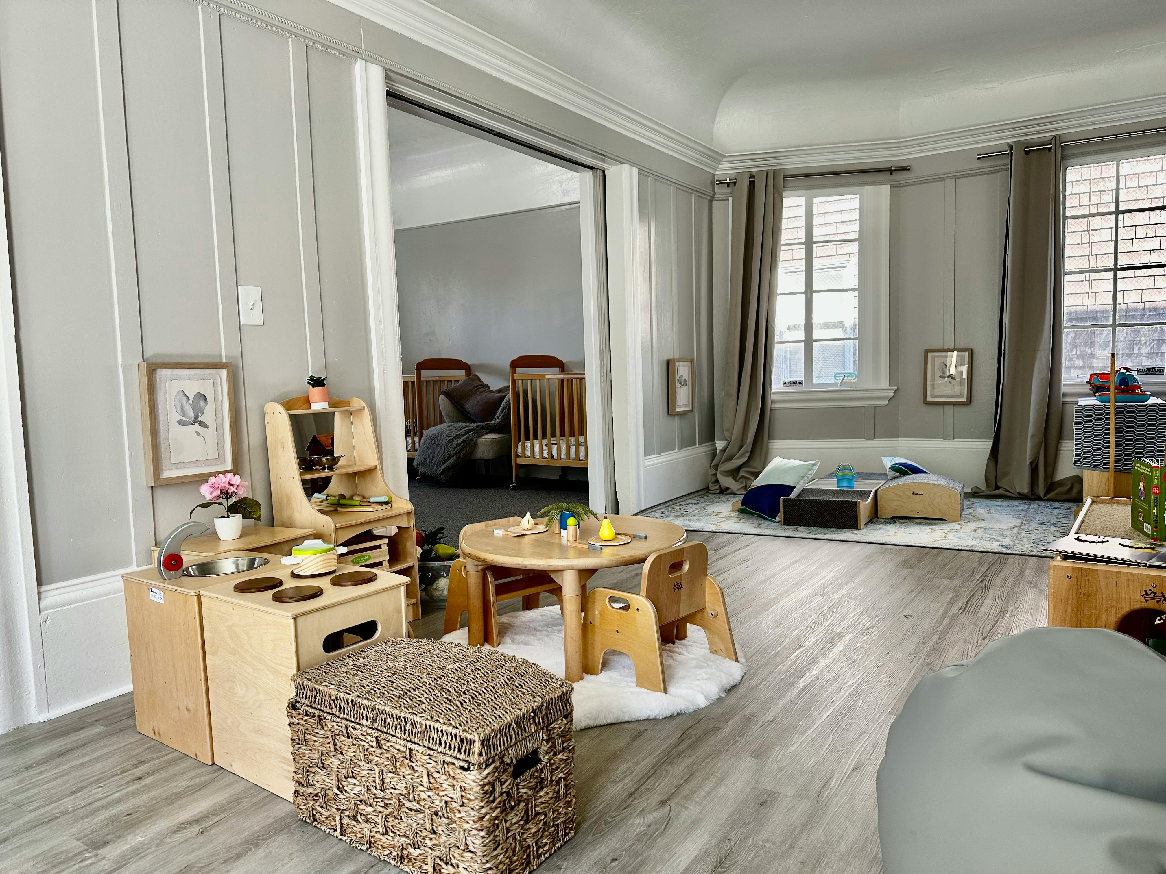 Playroom with child-sized table and chairs, play kitchen, and cribs in background