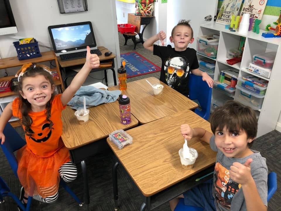 Three children sitting at desks, eating from bowls and giving thumbs up