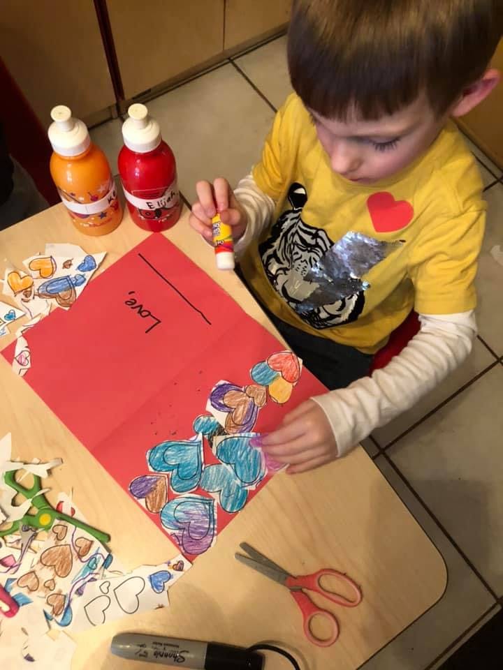 Child gluing paper hearts onto a red sheet of paper with "Love" written on it