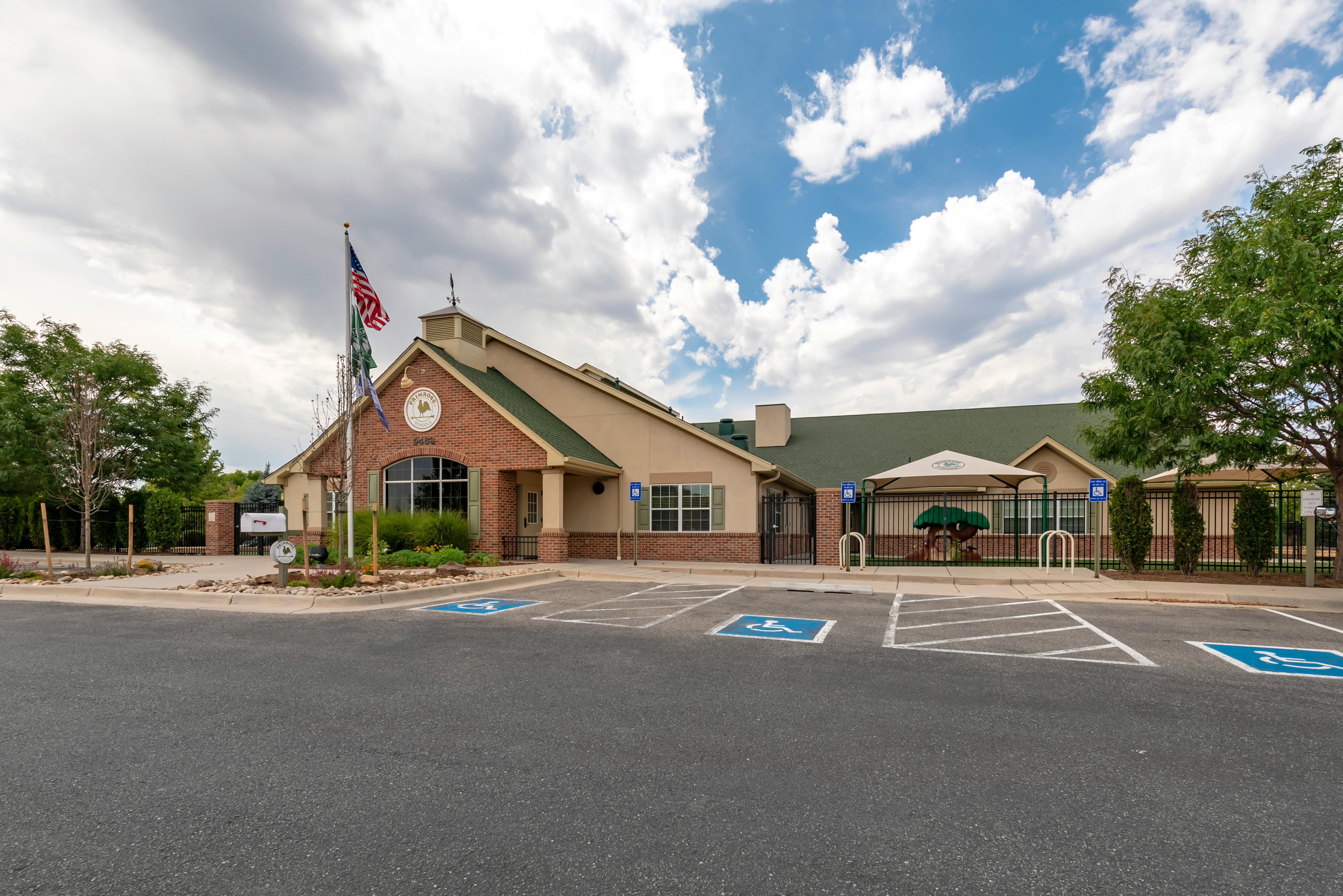 A beige and brick building with a green roof and flags outside, parking lot in front