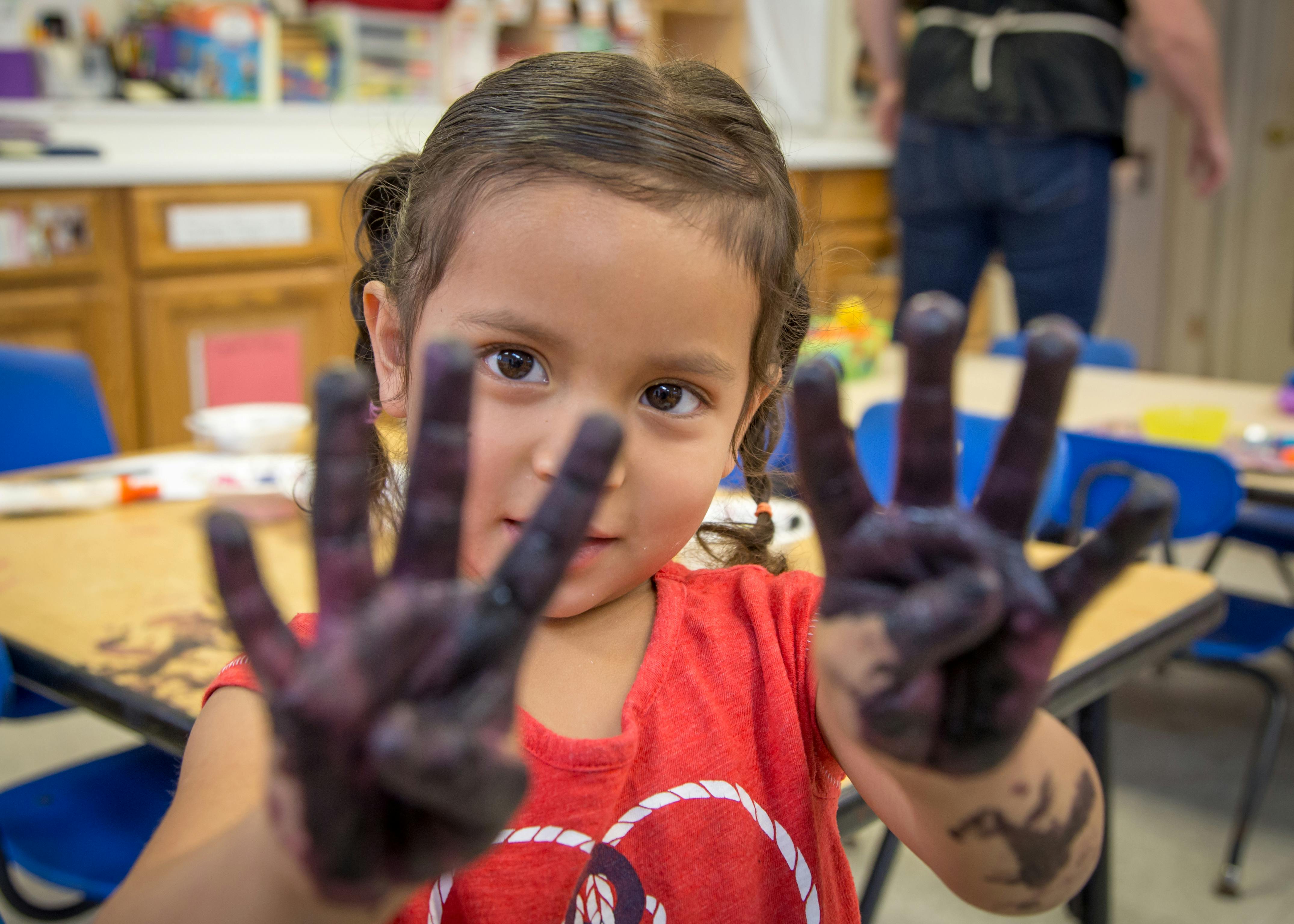 Child with painted hands holding up fingers in a classroom
