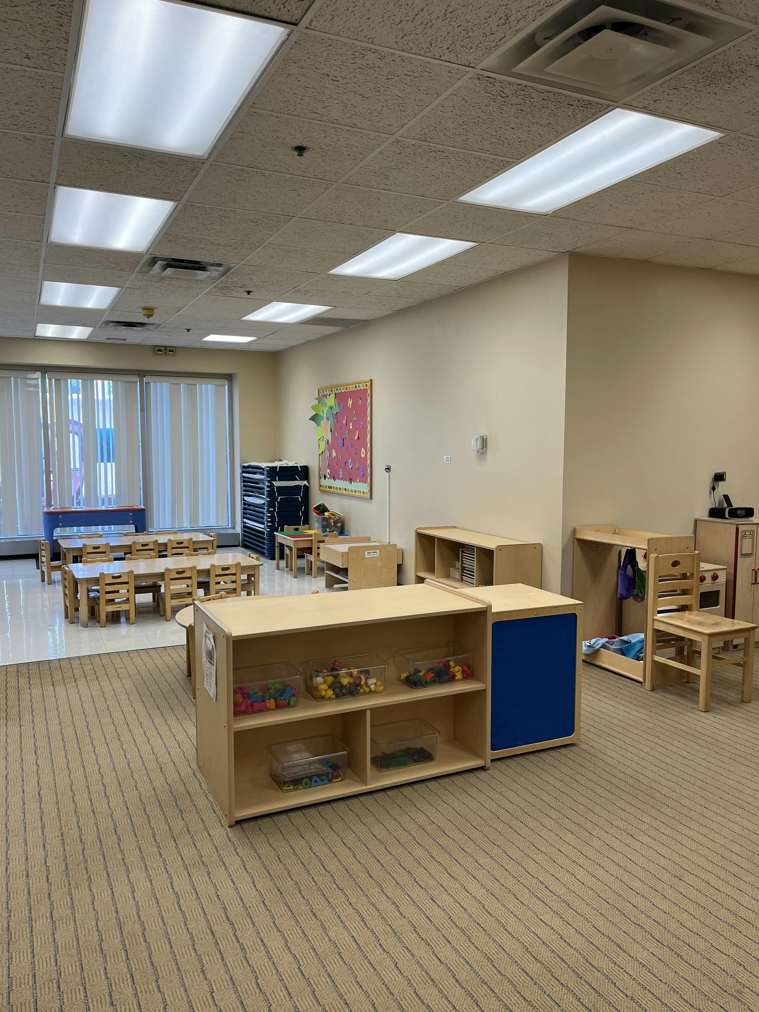An empty classroom with child-sized tables and chairs, shelves with toys, and windows