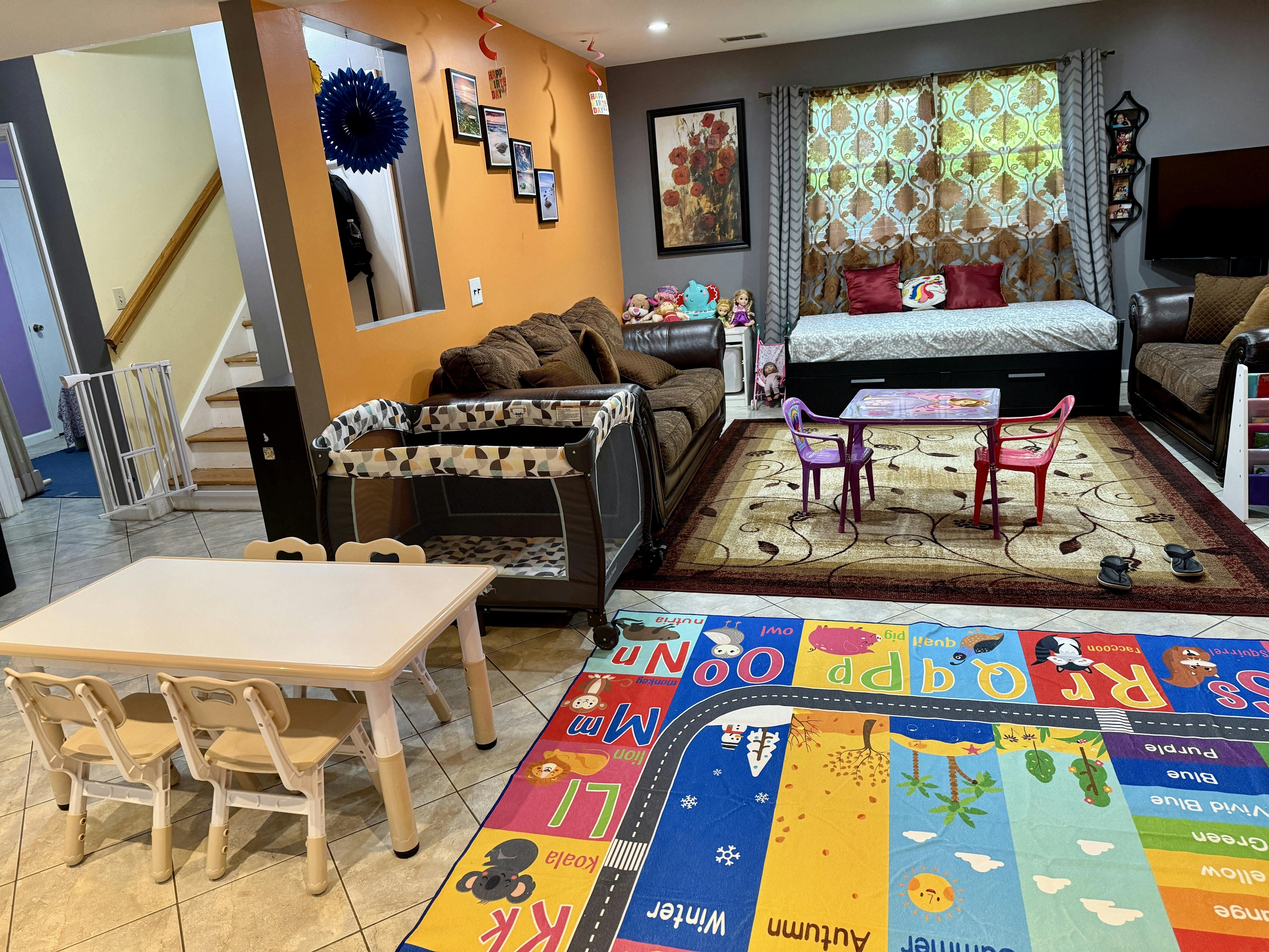 A living room with a playpen, children's tables and chairs, and a colorful alphabet rug