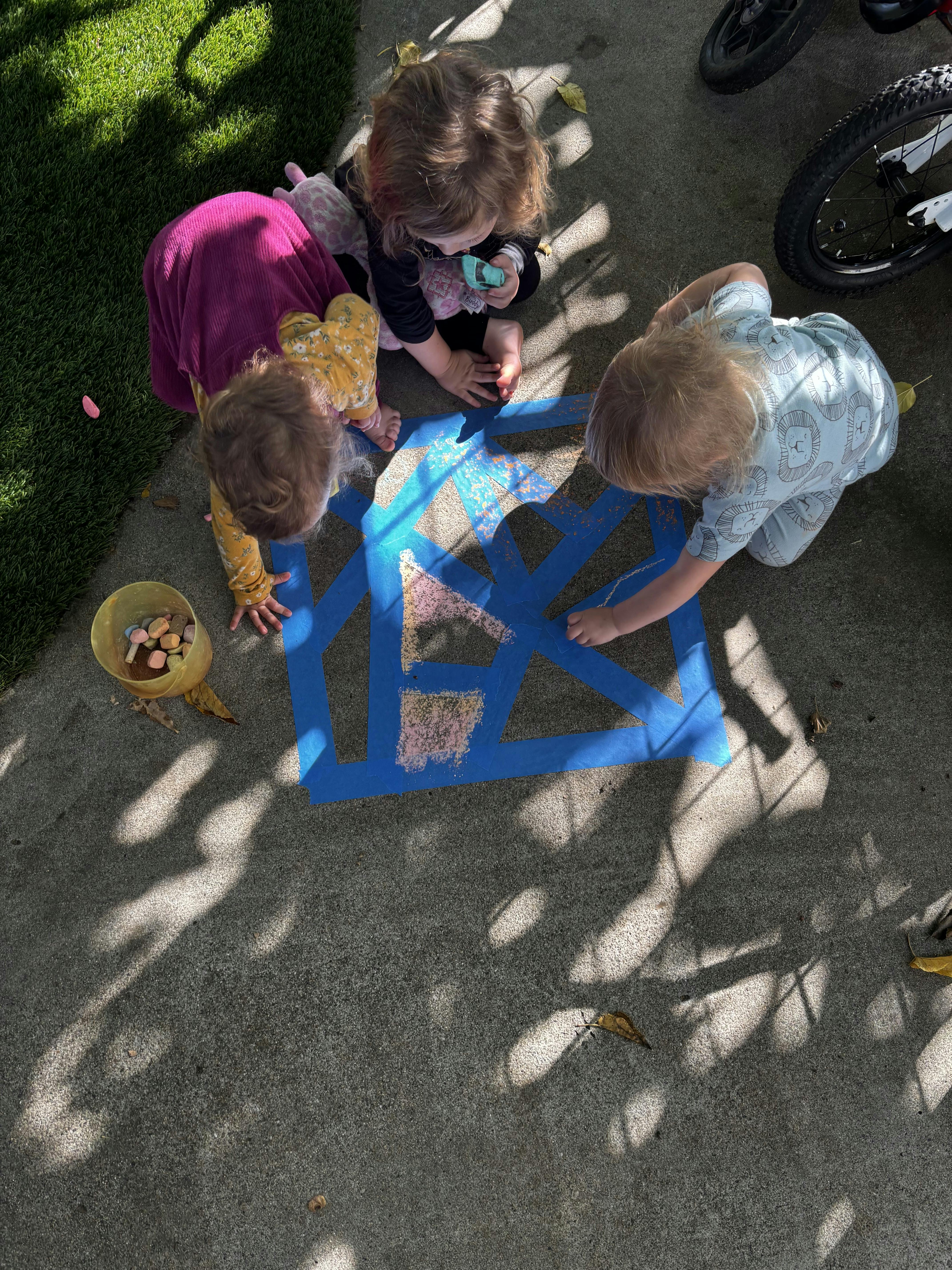 Three children drawing with chalk on concrete within a taped geometric pattern