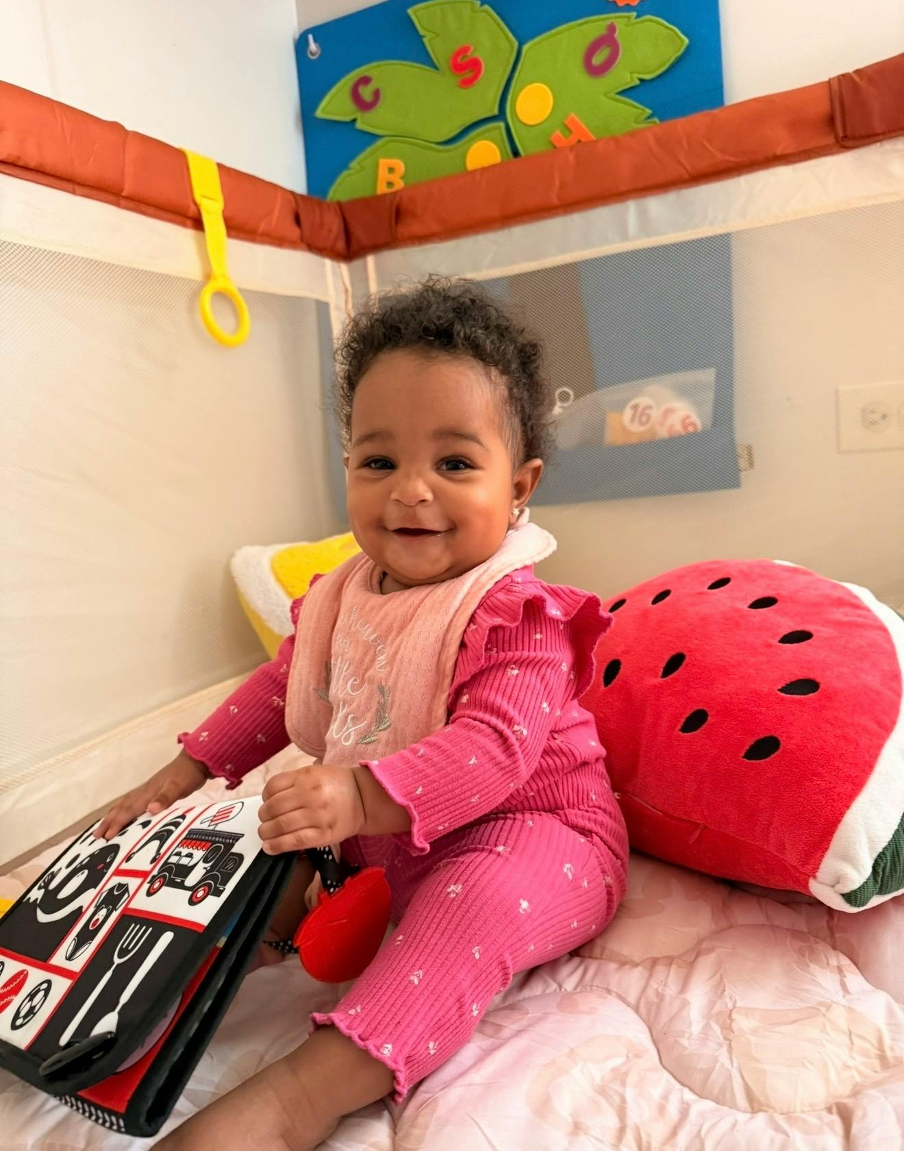 A smiling baby sitting in a playpen with a soft book and a watermelon pillow