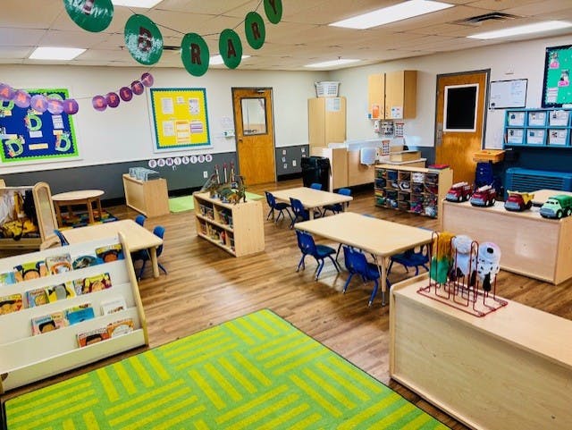 Classroom with small tables and chairs, shelves with toys and books, and a green rug