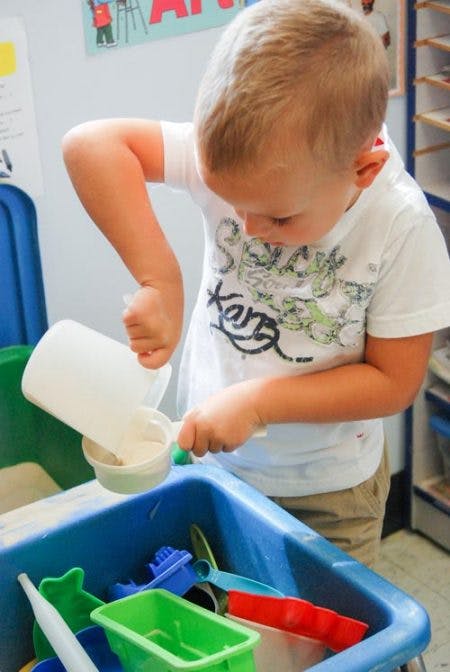 Child pouring from a white cup into another cup over a blue bin of toys